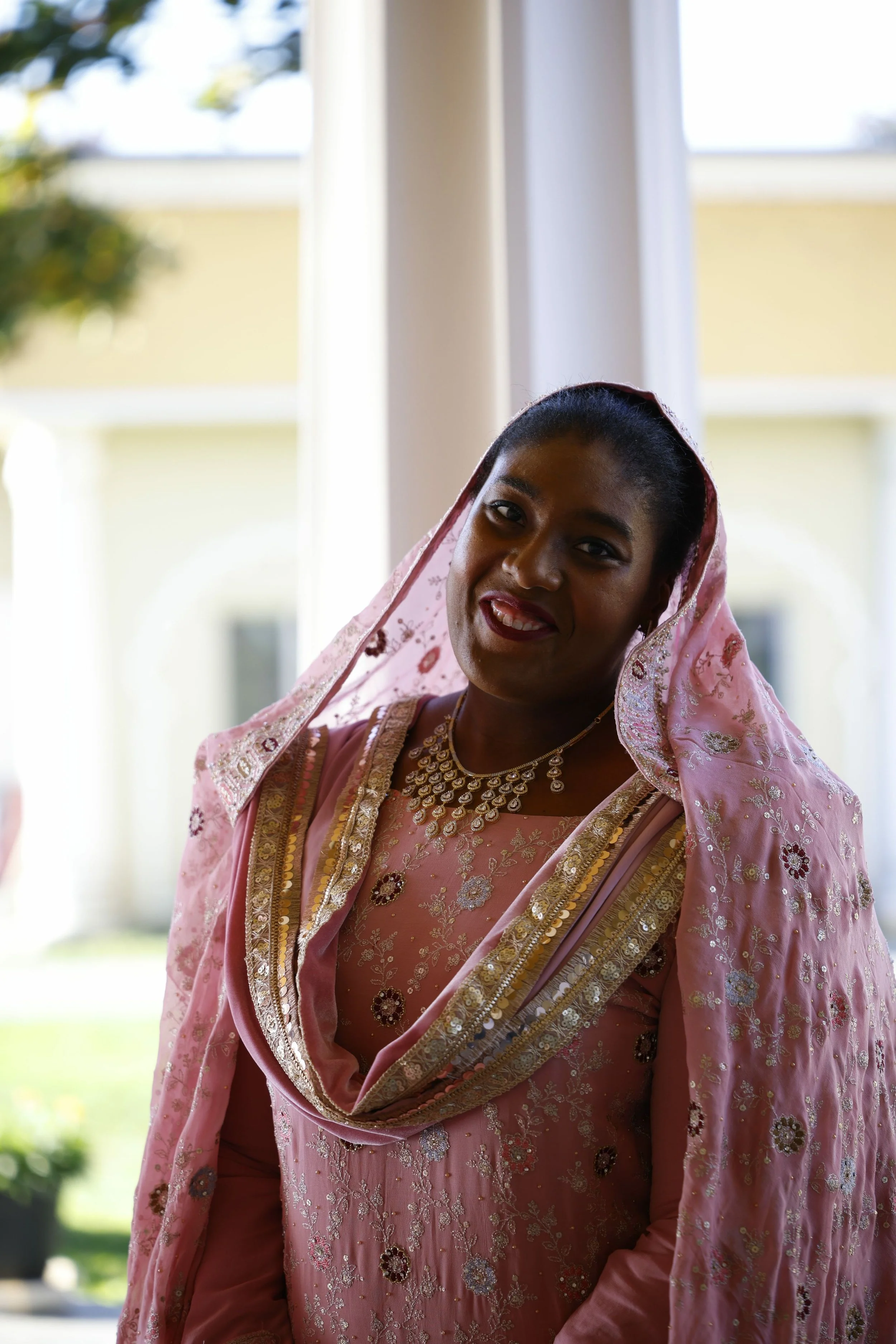 Woman in pink traditional dress with gold embroidery and jewelry, smiling indoors with sunlight and greenery visible outside.