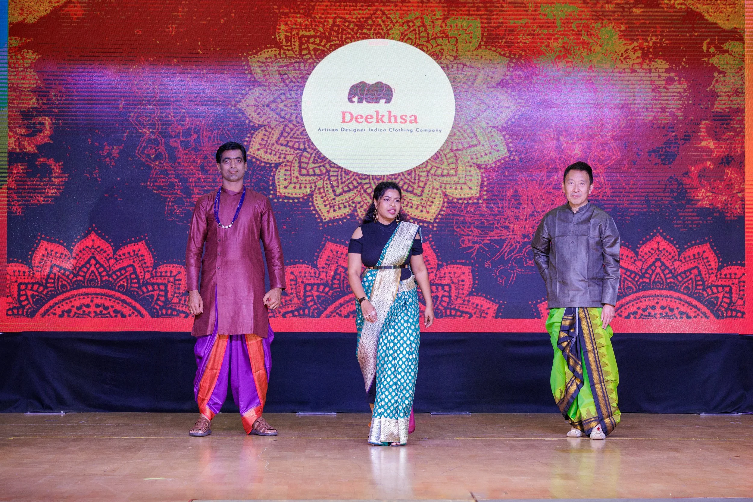 Three people in traditional Indian attire standing on a stage in front of a vibrant red and purple background with a circular logo that reads 'Deeksha - Artisan Designer Indian Clothing Company'.
