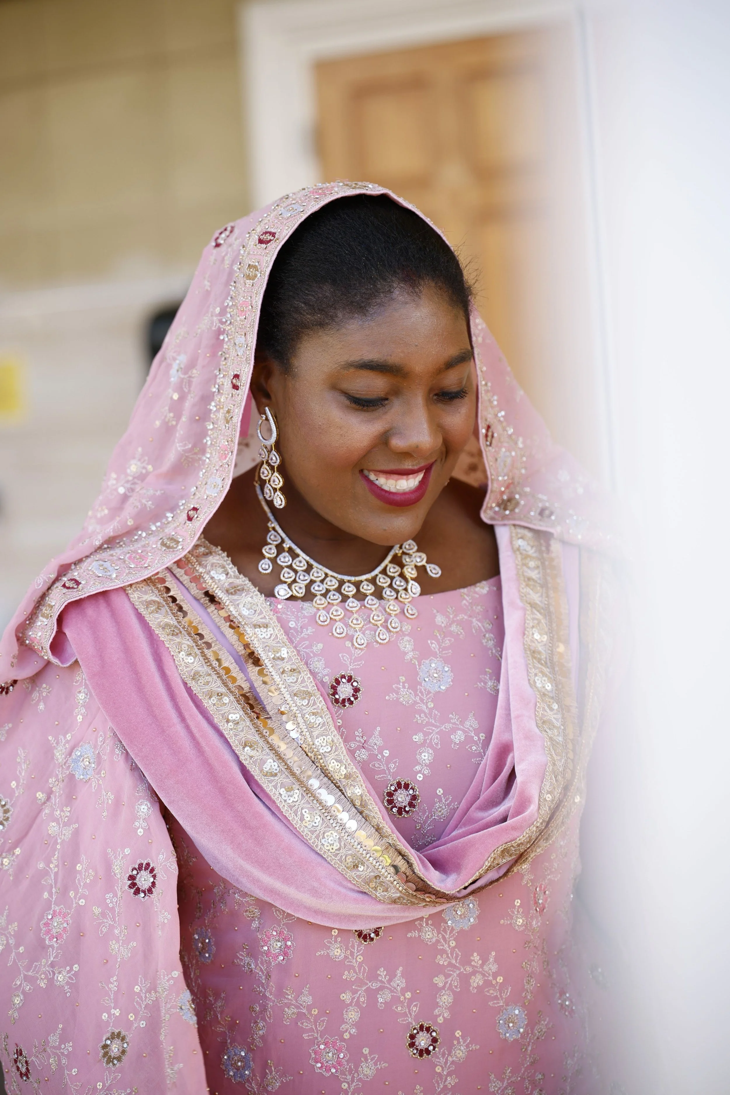 A woman in a pink, embroidered traditional dress and jewelry, smiling with her eyes closed, standing near a white wall.