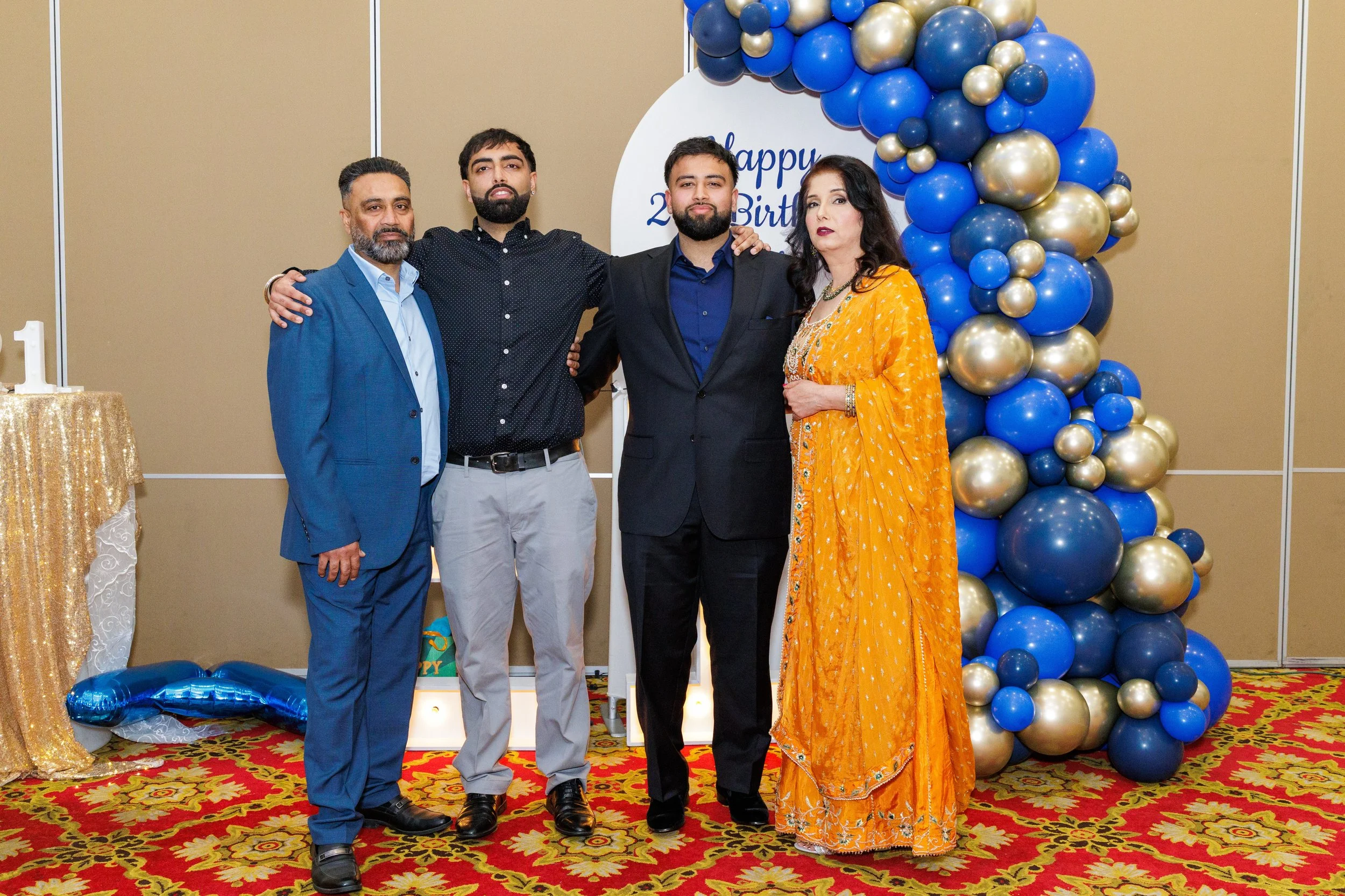Four people standing together at a birthday celebration, with a backdrop of balloons in blue, gold, and silver, and a sign that says 'Happy 2nd Birthday'. One man is wearing a blue suit, another a black shirt, the third a black suit, and a woman is d