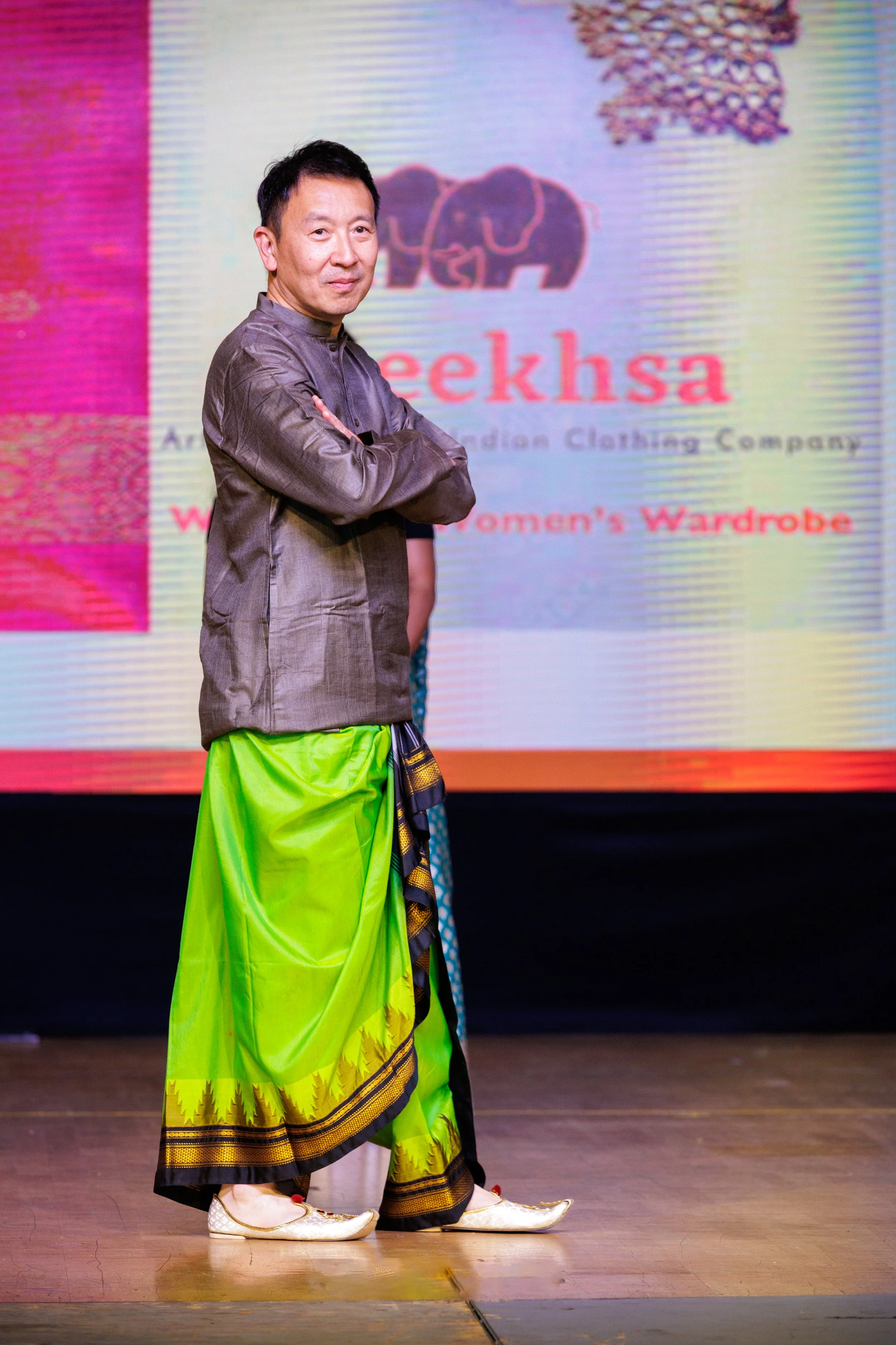 A man standing on stage in traditional attire, wearing a grey jacket and a bright green and black dhoti, with a woman in traditional clothing in the background. Behind them, a large screen displays a logo with an elephant and the text 'Geekhsa' and p