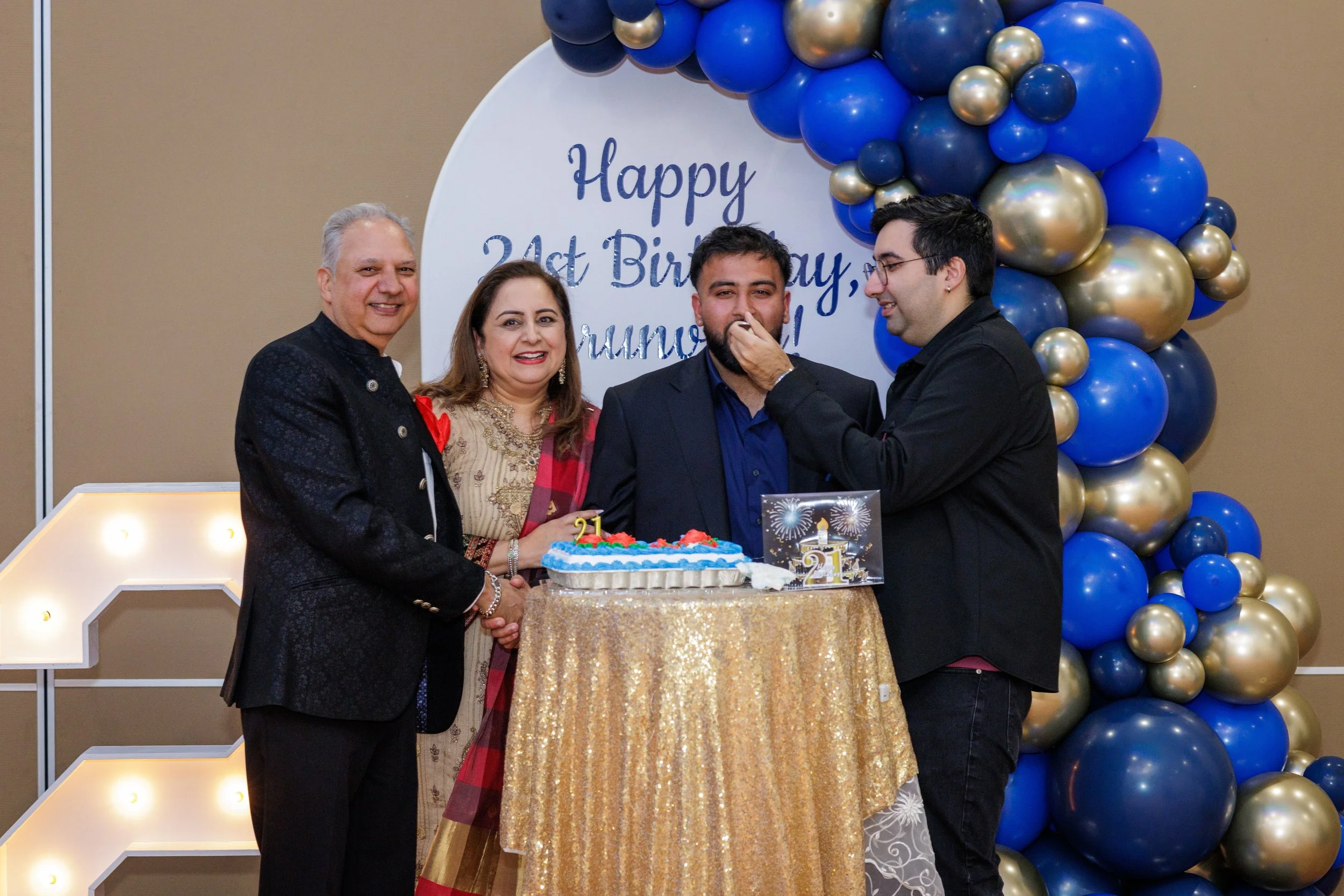 Group of four people celebrating a birthday with a cake, gold and blue balloons, and a 'Happy 21st Birthday' sign in the background.
