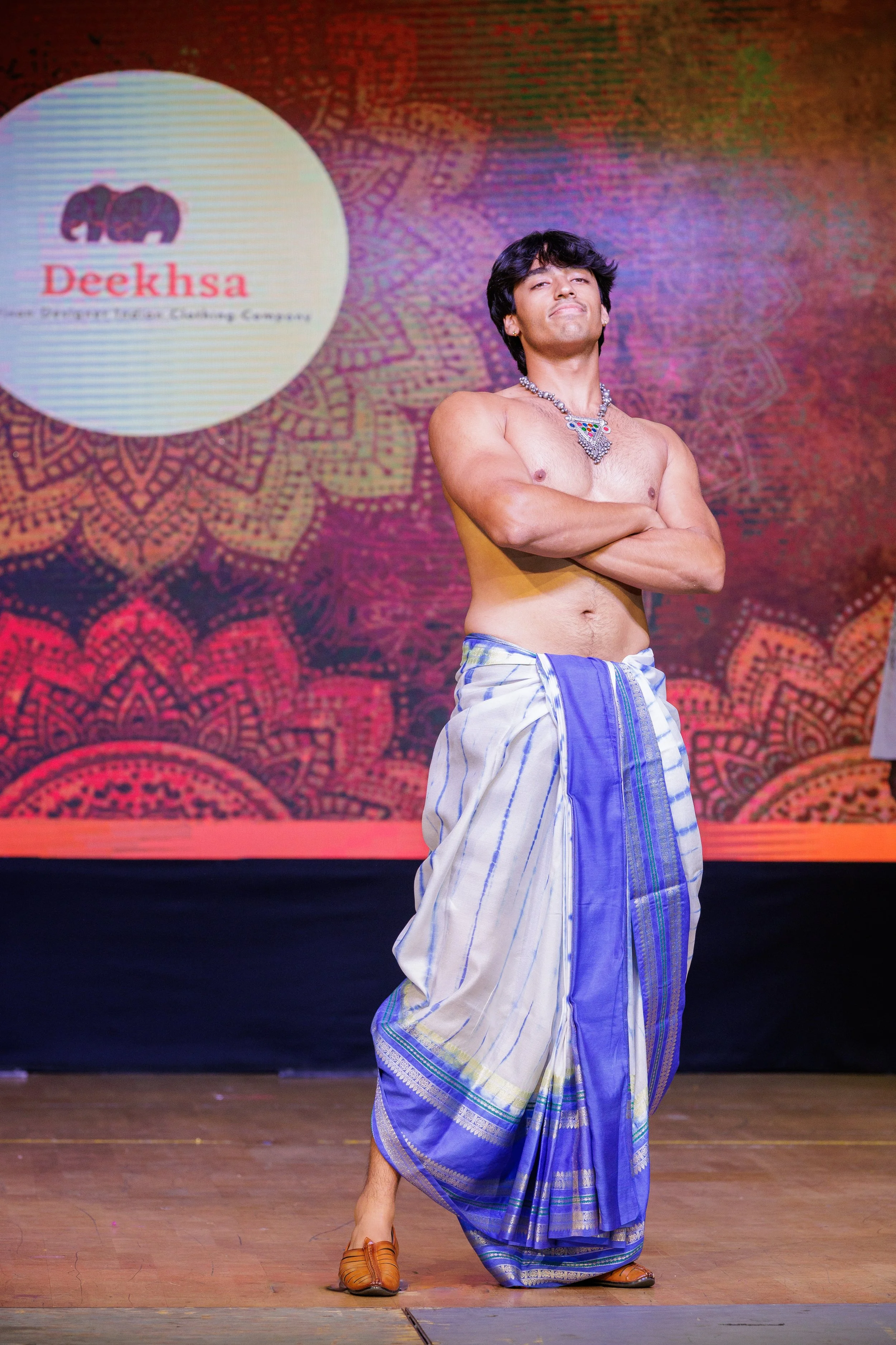 A man in traditional Indian attire standing on stage with arms crossed, shirtless, wearing a dhoti and sandals, against a colorful background with a logo that reads "Deeksha".