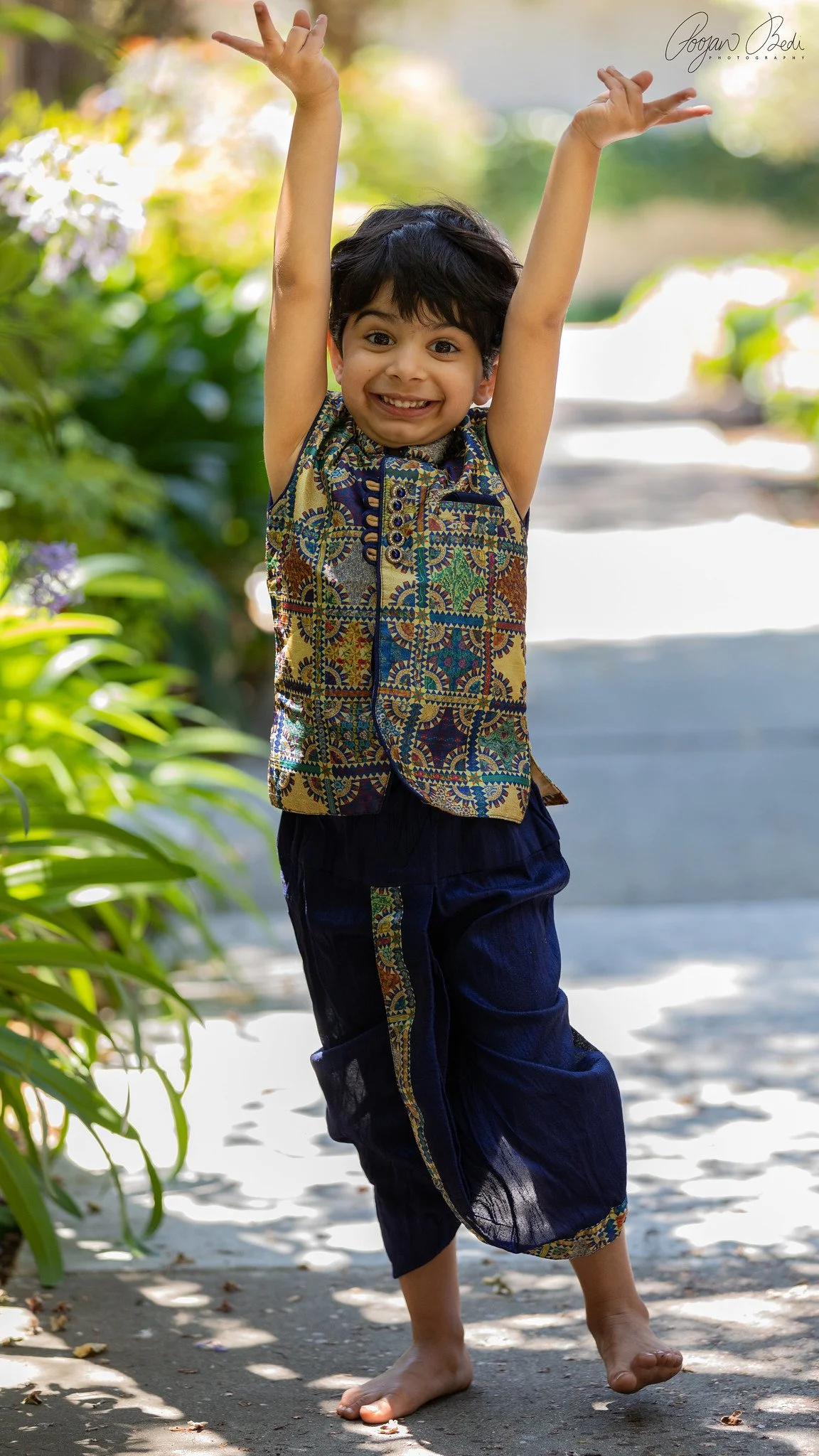 Happy young boy with short dark hair smiling and raising his arms barefoot outdoors in a garden or park.