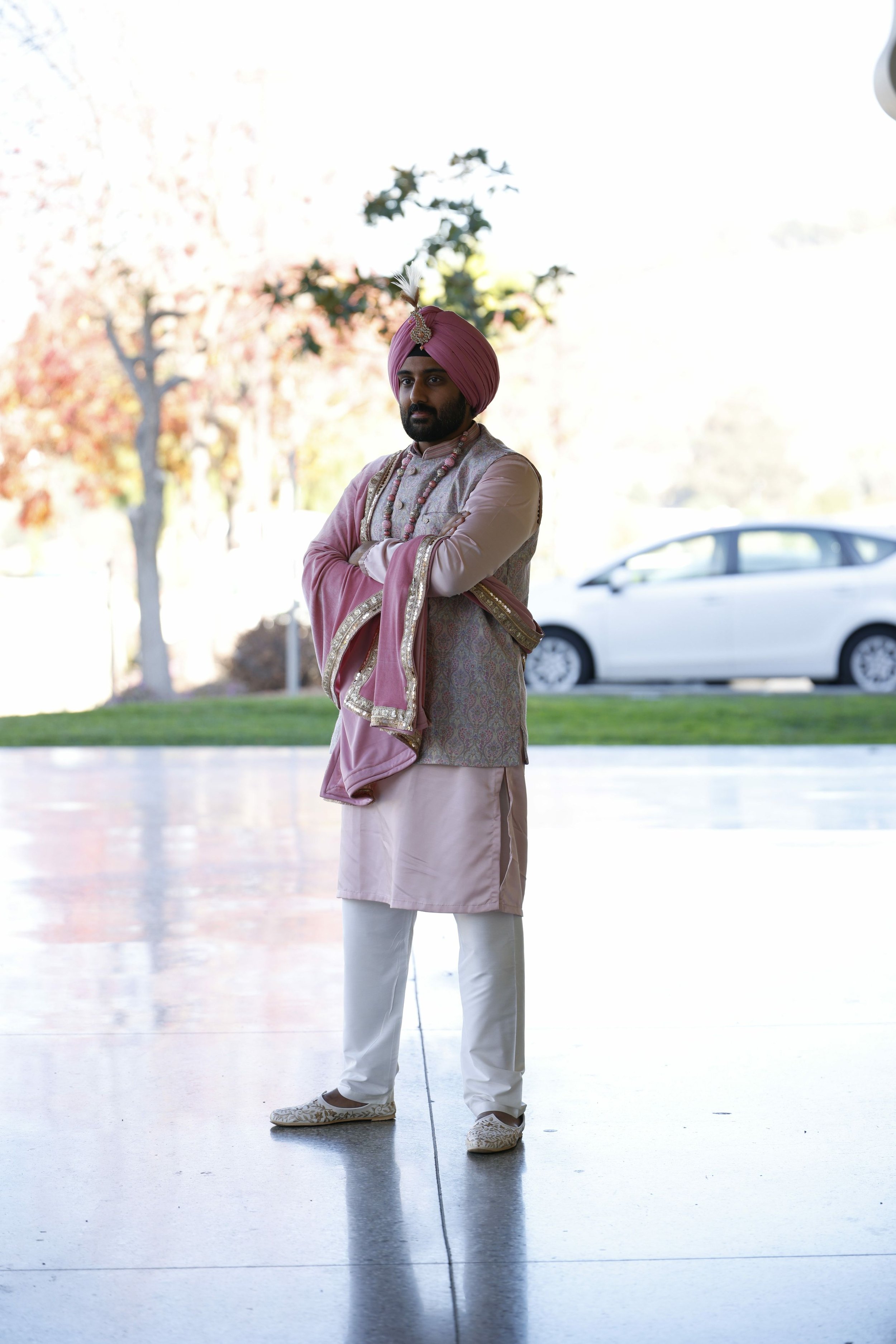 A man dressed in traditional Indian attire, including a pink turban with a feather, standing outdoors with arms crossed, a car in the background, and autumn trees.