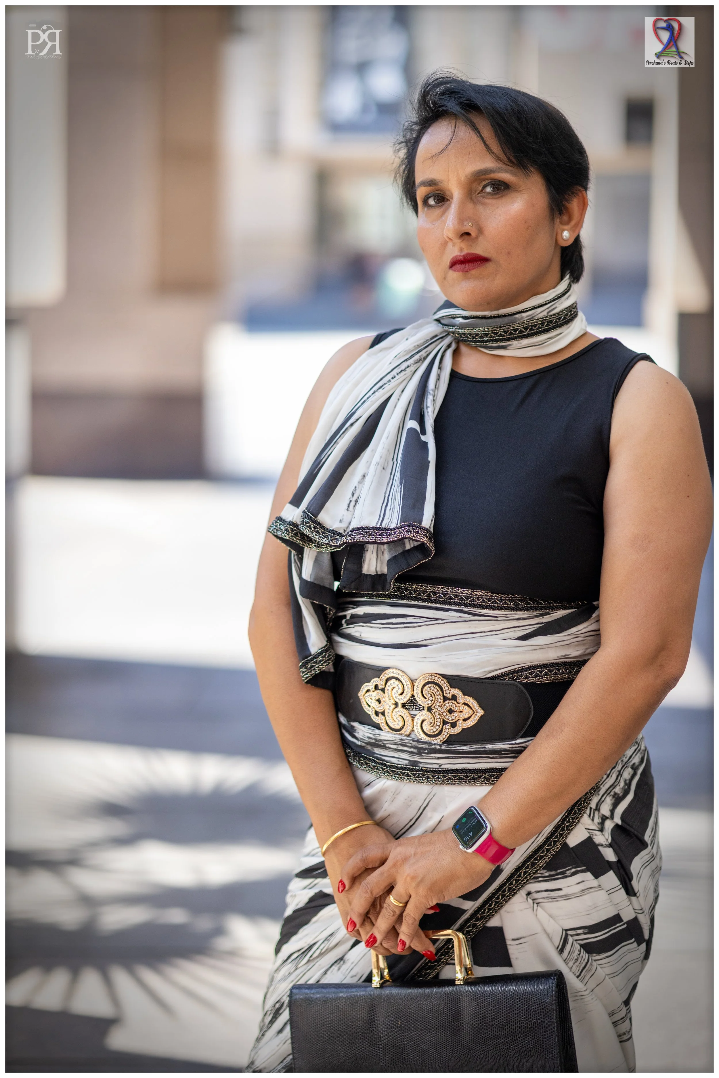 A woman with short black hair, dressed in a black top and a black-and-white patterned skirt with a matching scarf, holding a black handbag. She is also wearing a smartwatch and jewelry, standing outdoors with a blurred background.
