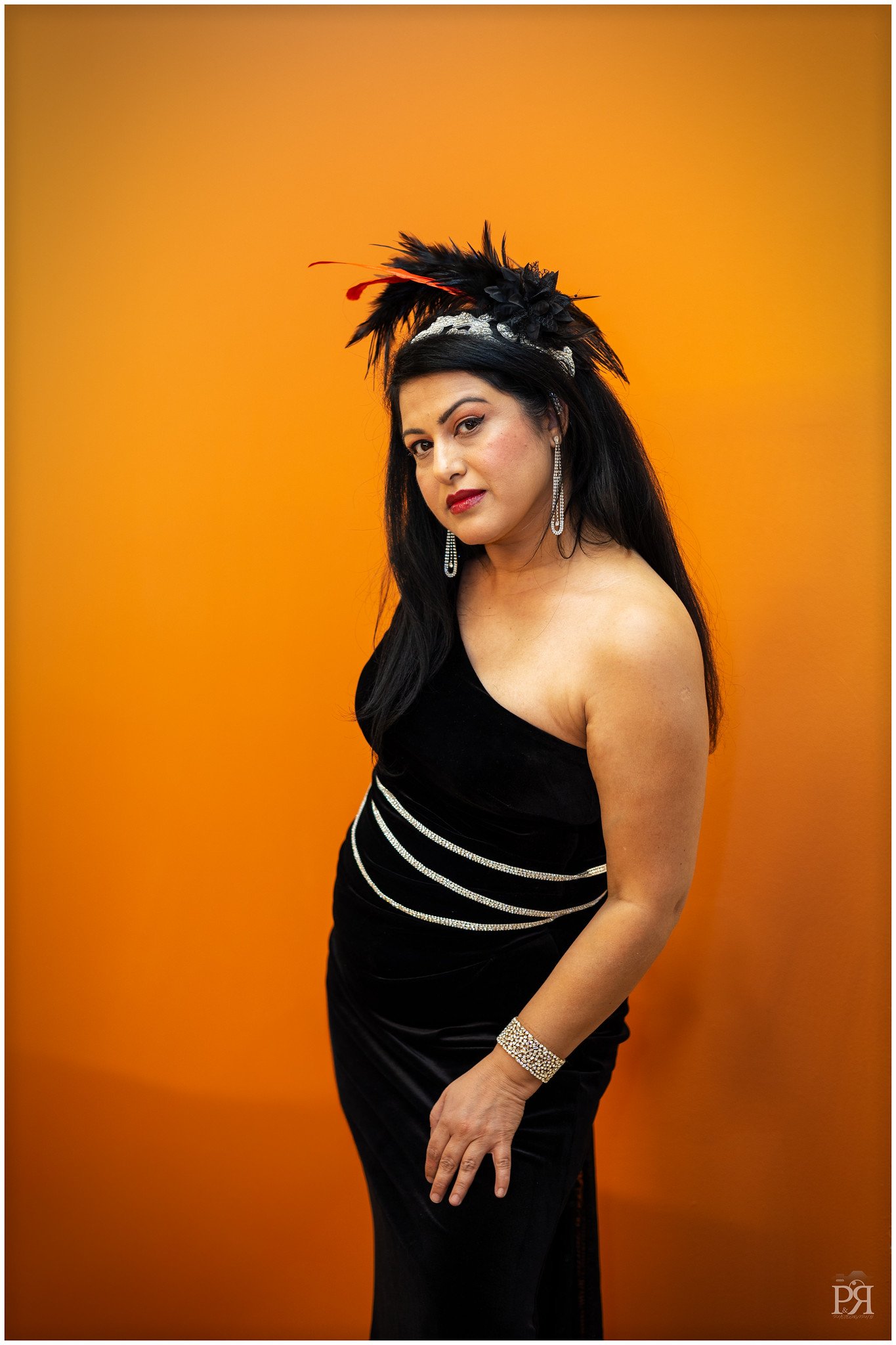 A woman in a black evening gown with silver embellishments, posed against an orange background, wearing a feathered headpiece and jewelry.