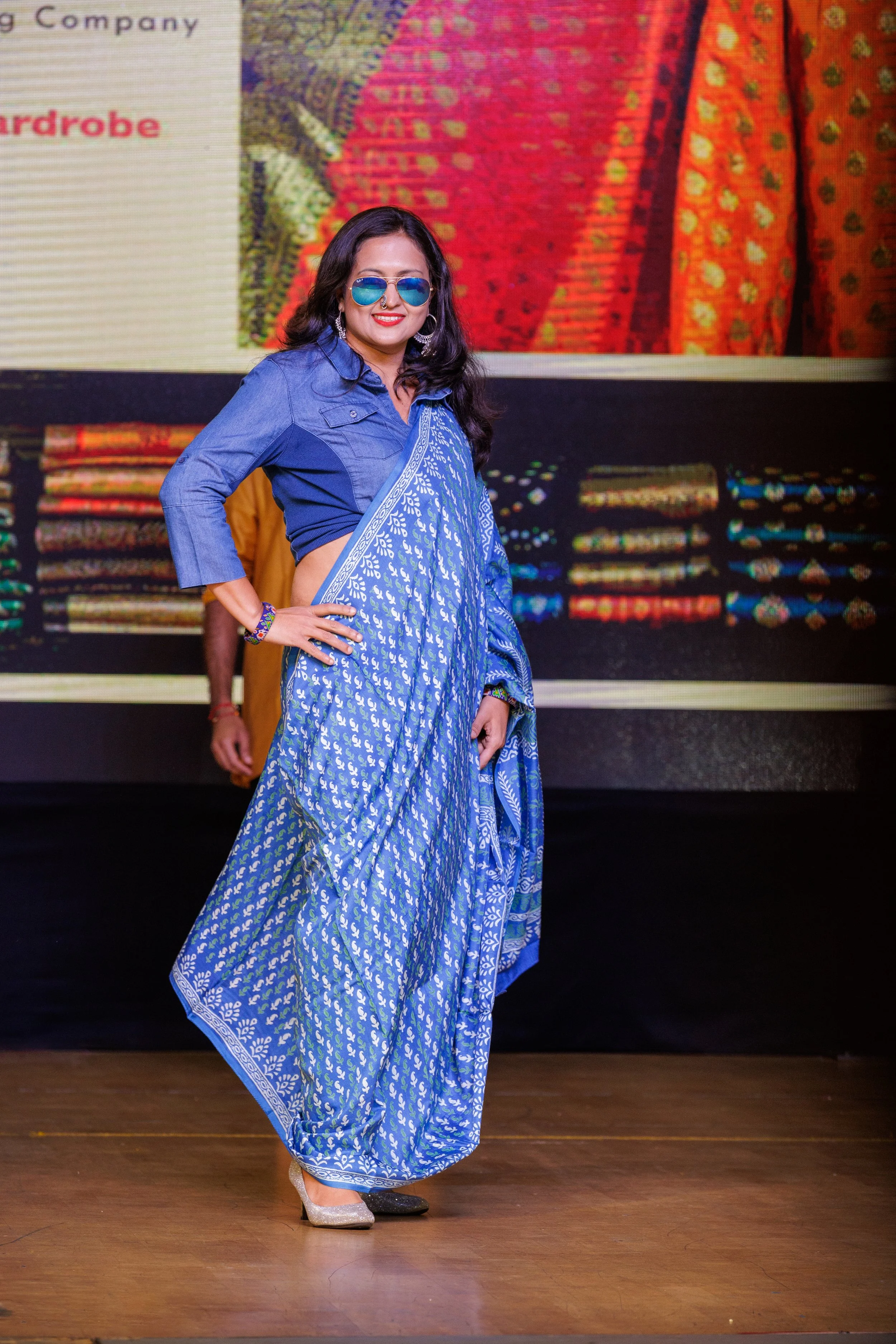 Woman in a blue saree with a denim shirt and sunglasses posing on stage at a cultural event.