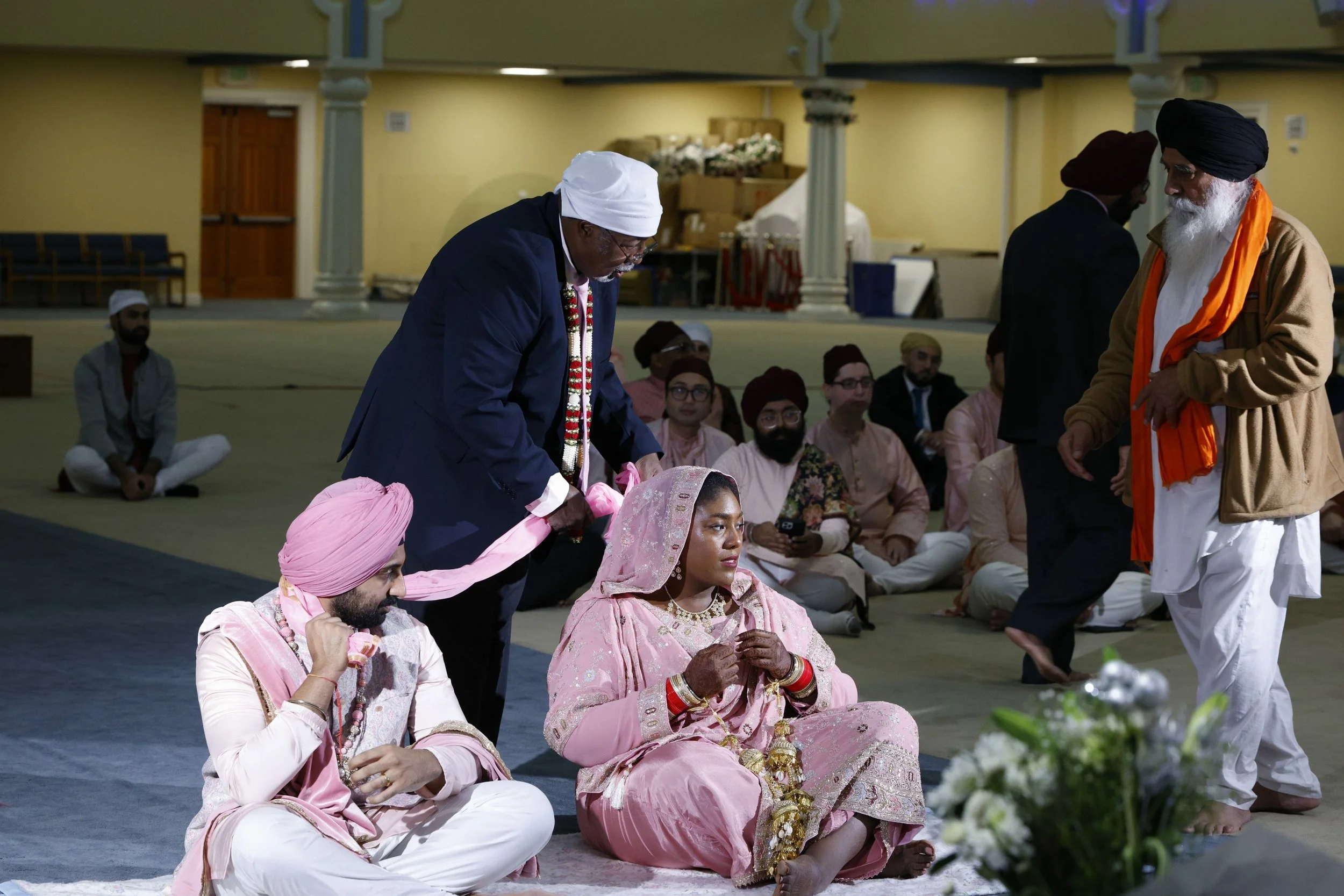 Indian wedding ceremony with a bride in a pink traditional outfit and woman holding jewelry, surrounded by men in traditional and formal attire, seated on the floor in an indoor hall.