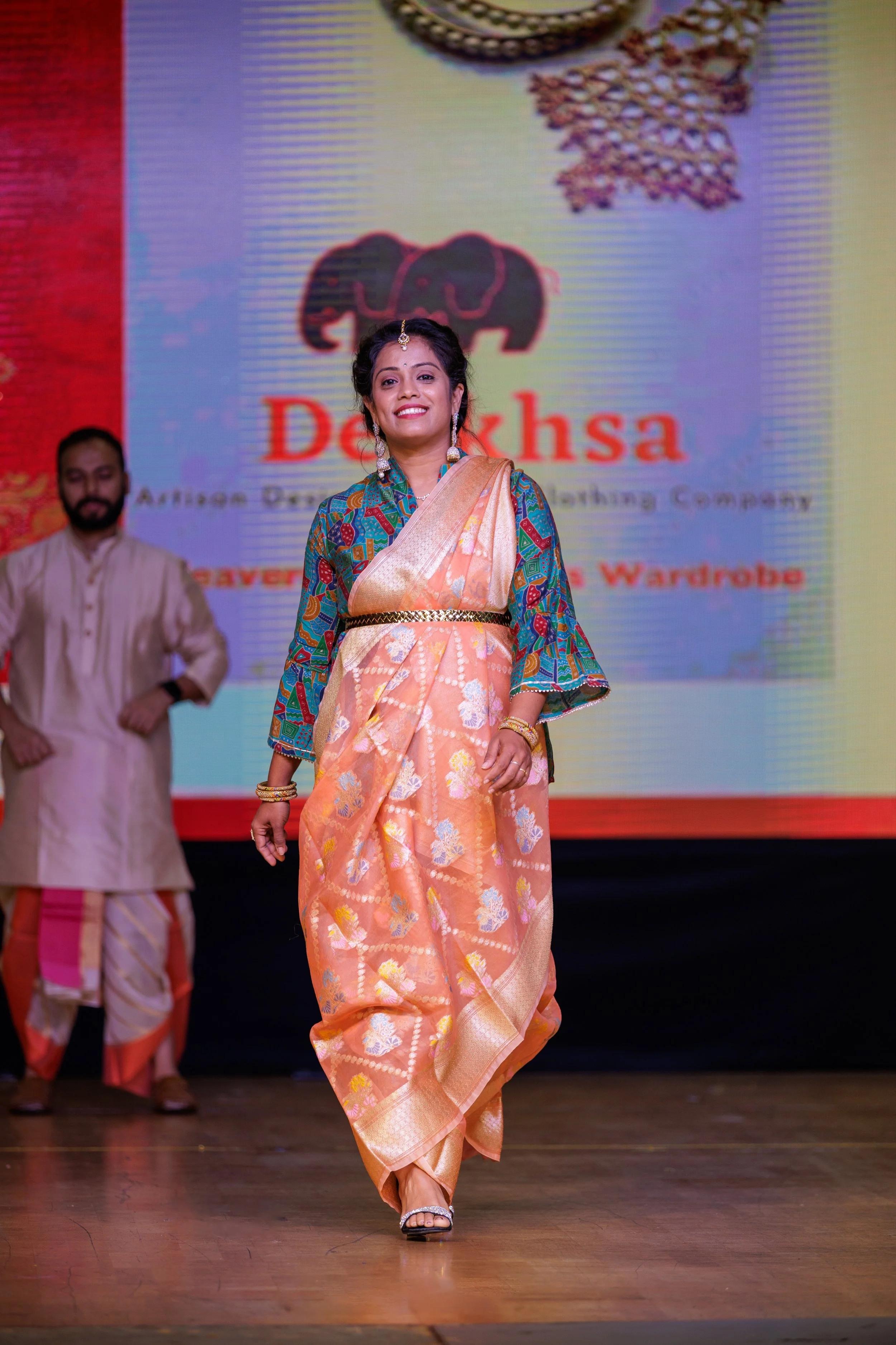 Woman walking on stage wearing a traditional saree with a golden belt, colorful blouse, and jewelry. Background displays the logo and the name "Deeksha" and other text, with a man standing behind her.