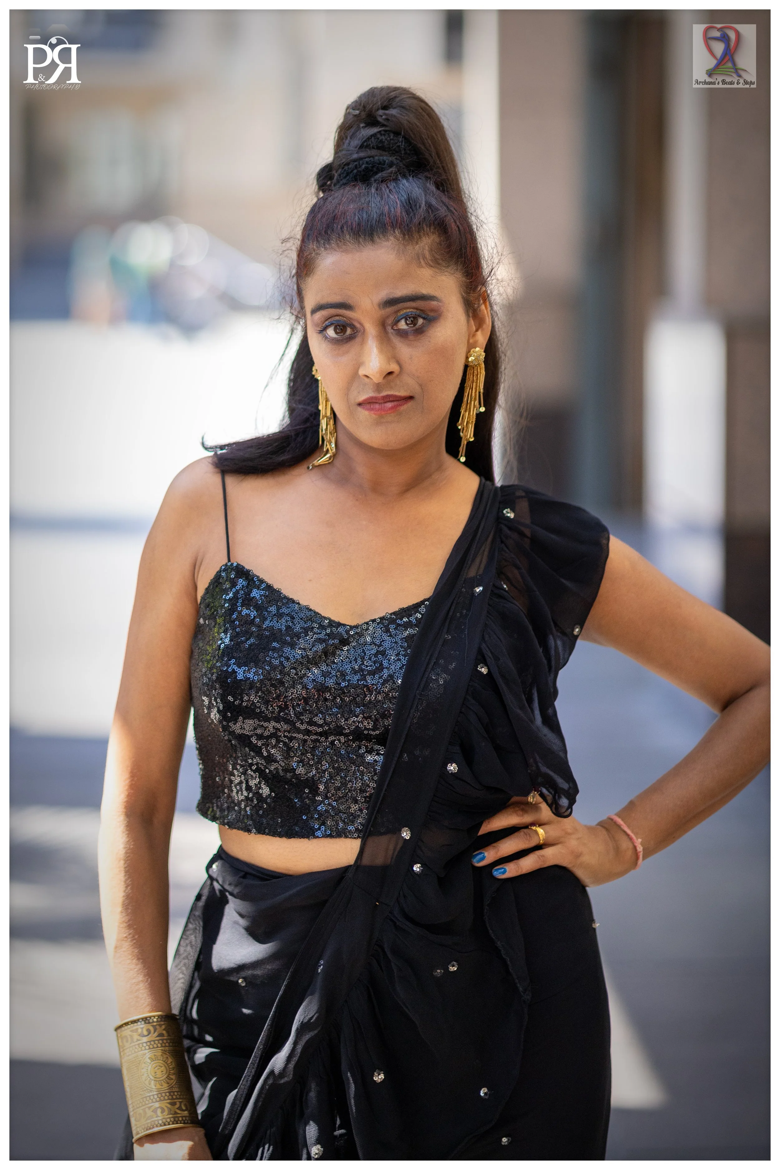 A woman wearing a black and sequined crop top and sari with gold jewelry, standing with her hand on her hip on a city street.