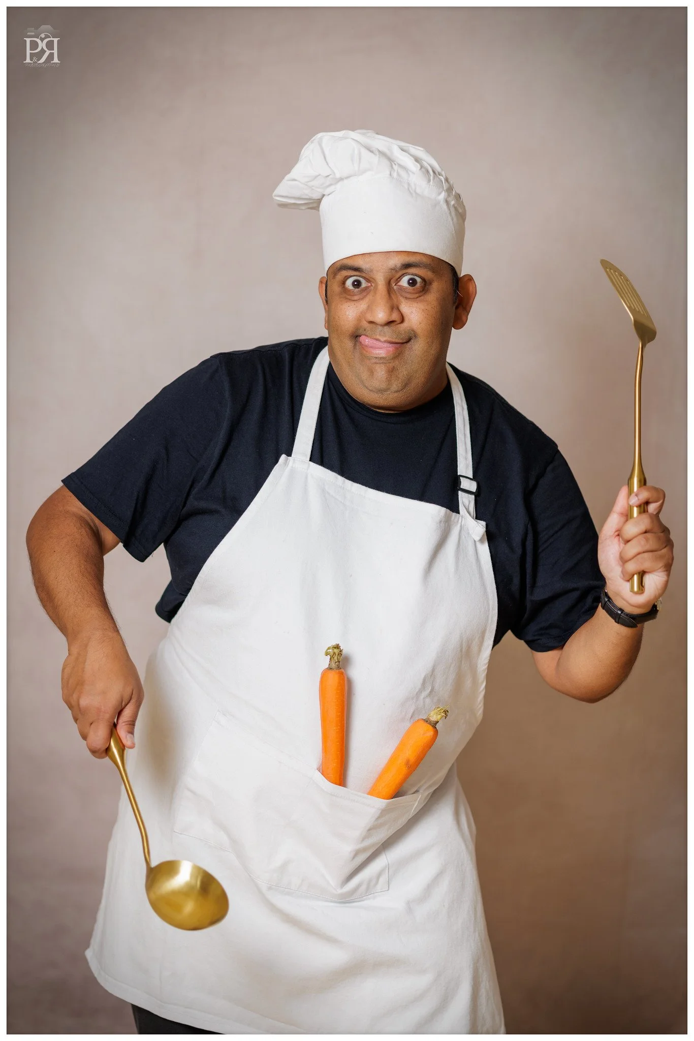 A man dressed as a chef wearing a white apron and chef's hat holding a gold serving spoon and fork, with two carrots in his apron pocket, standing against a plain neutral background.