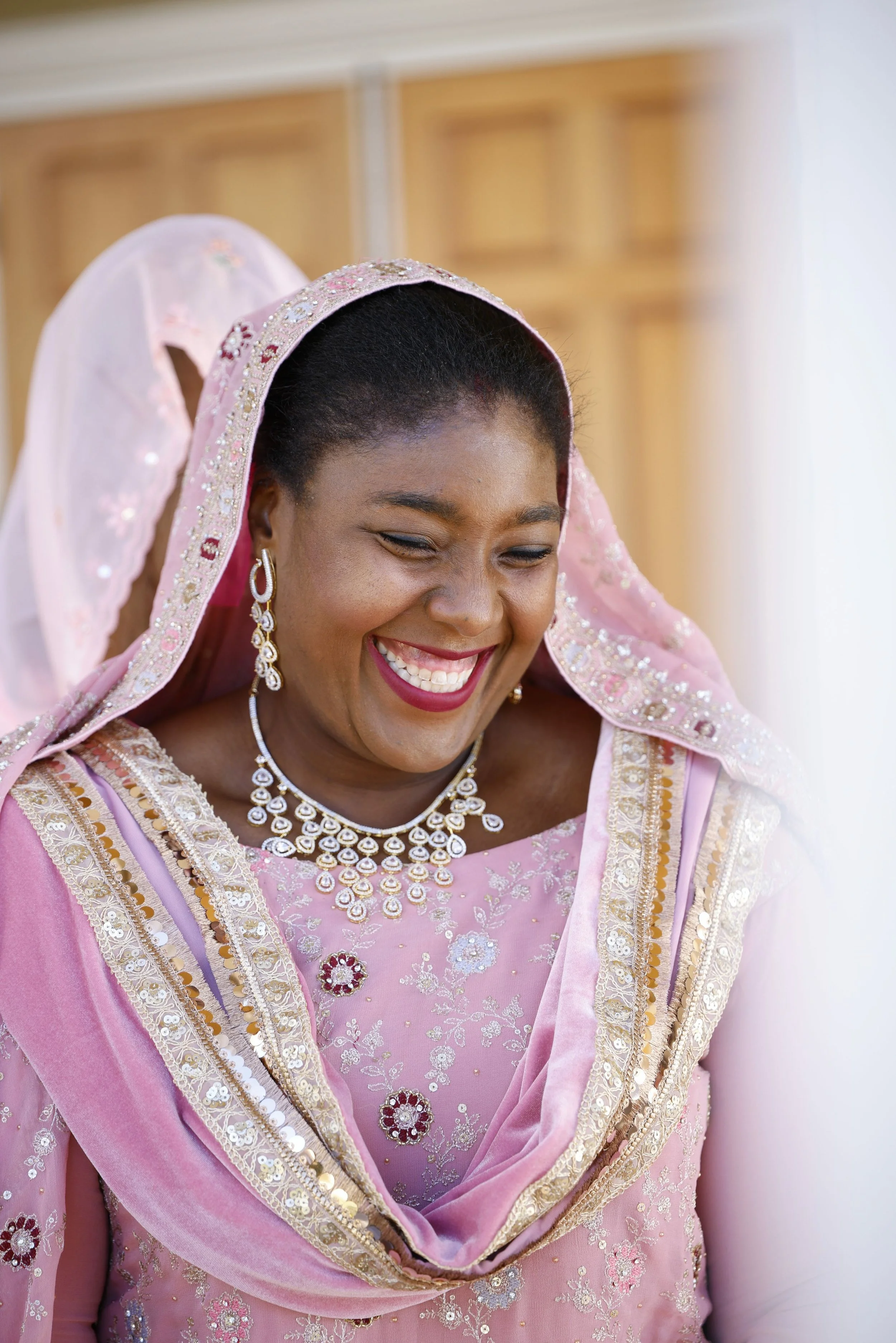 A woman dressed in pink traditional attire with jewelry, smiling and looking down.