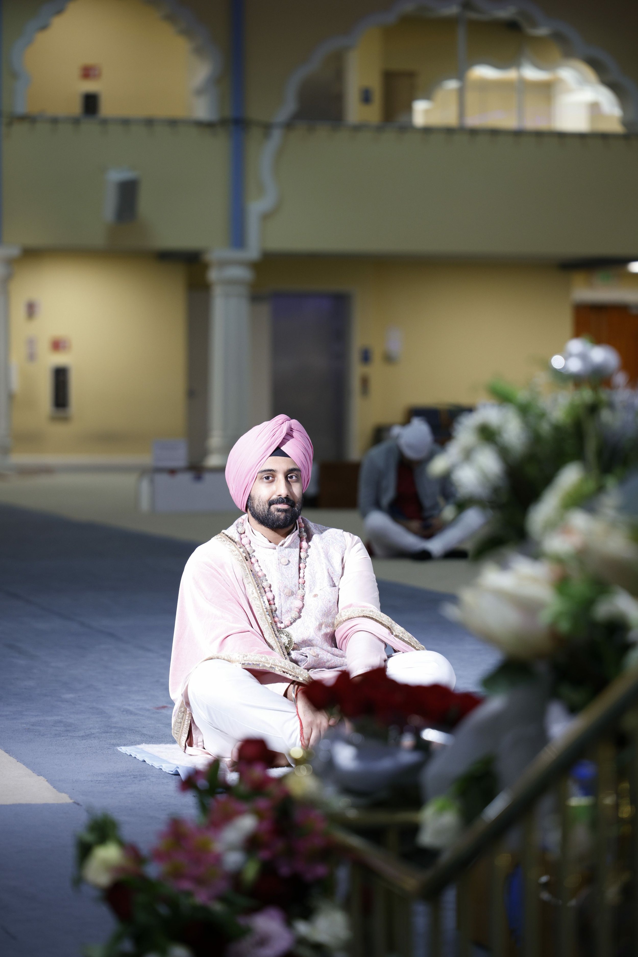 A man dressed in traditional pink and white Indian attire, including a turban and jewelry, sitting on the floor during a ceremonial event, with floral arrangements in the foreground and a decorated indoor setting in the background.