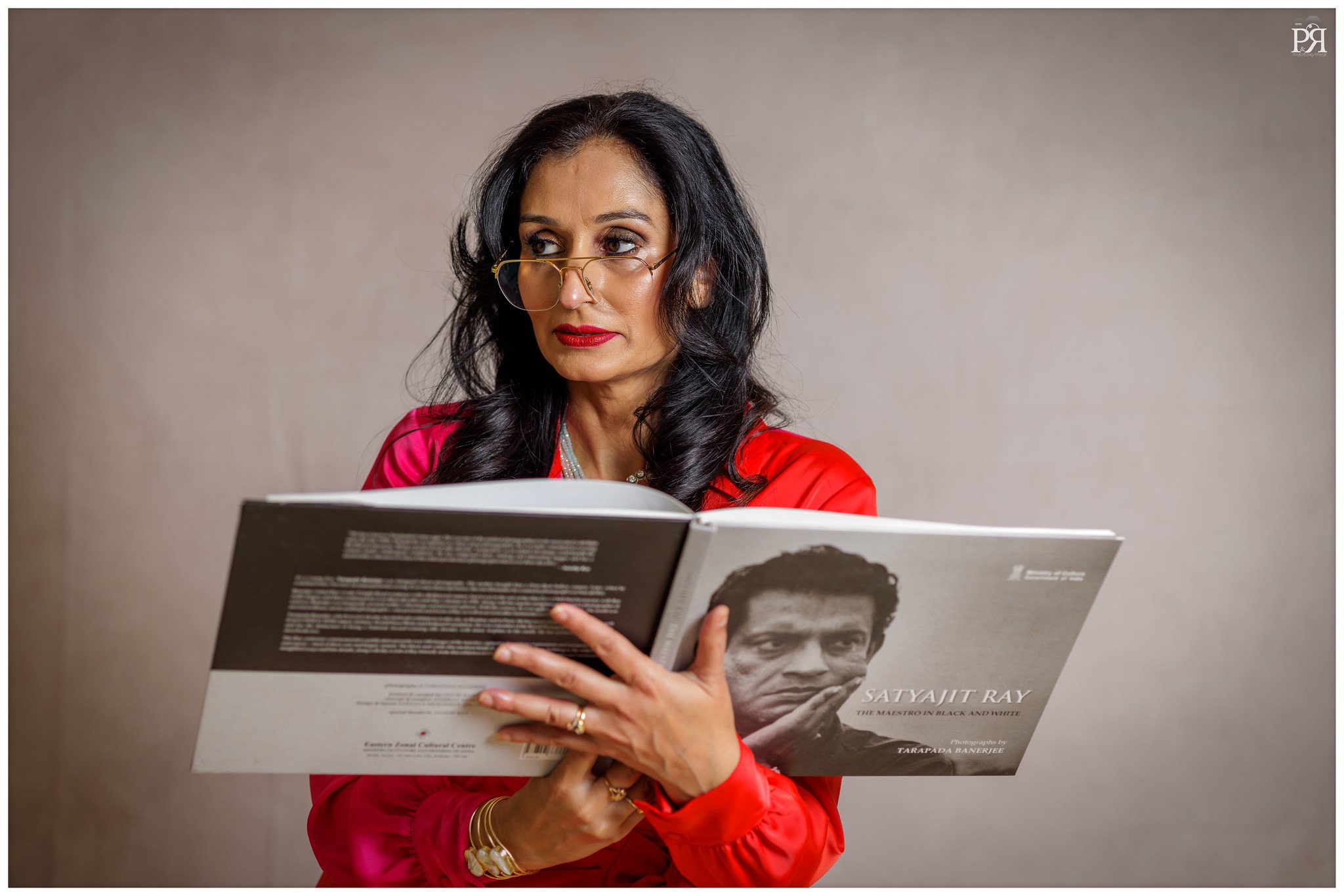 A woman with dark wavy hair, red lipstick, and glasses is reading a book titled 'Satya Jit Ray: The Maestro in Black and White' by Tarapada Banerjee, in front of a plain beige wall.