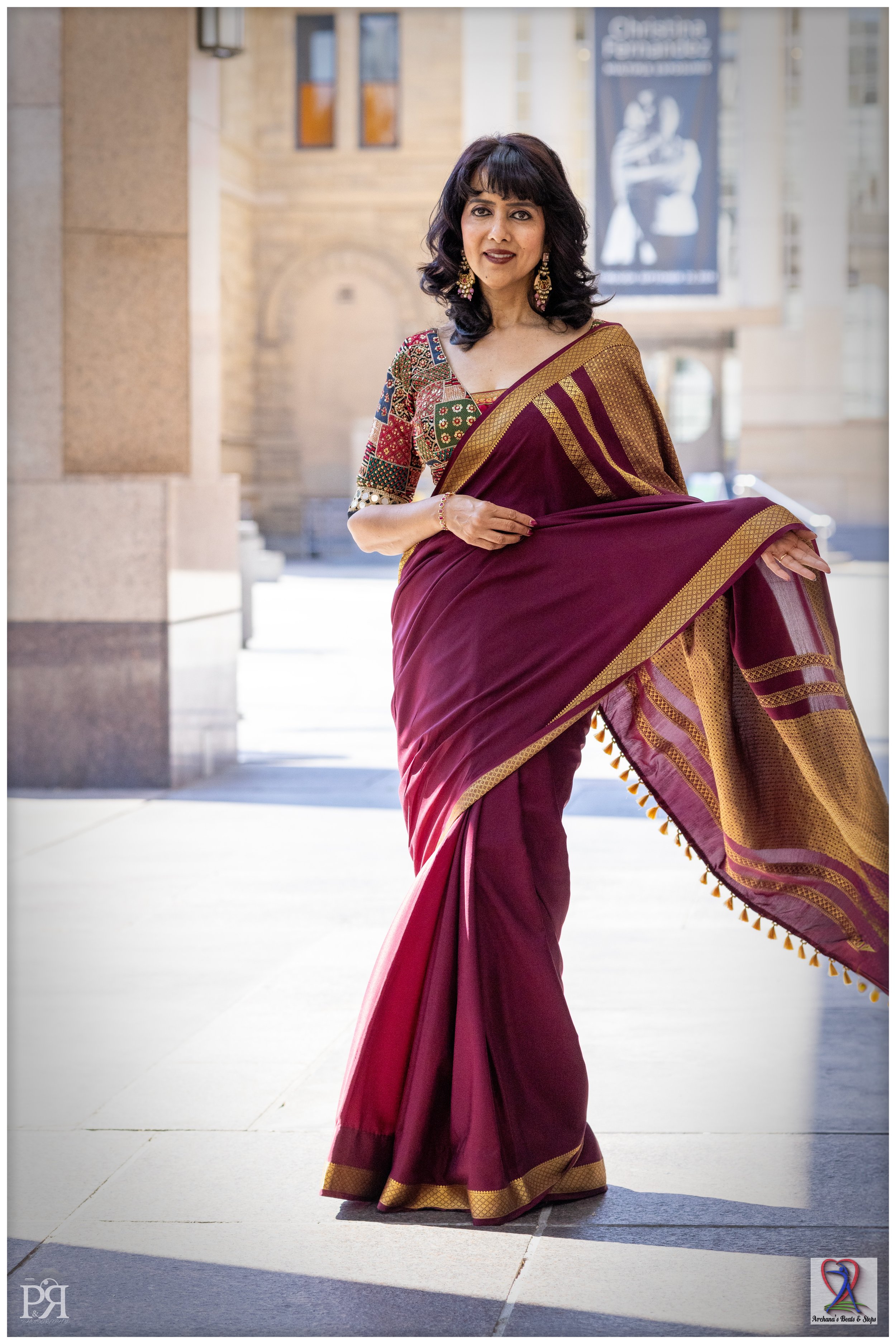 A woman dressed in a traditional maroon saree with a gold border, standing outdoors in front of a historic building, smiling and holding her saree with one hand.