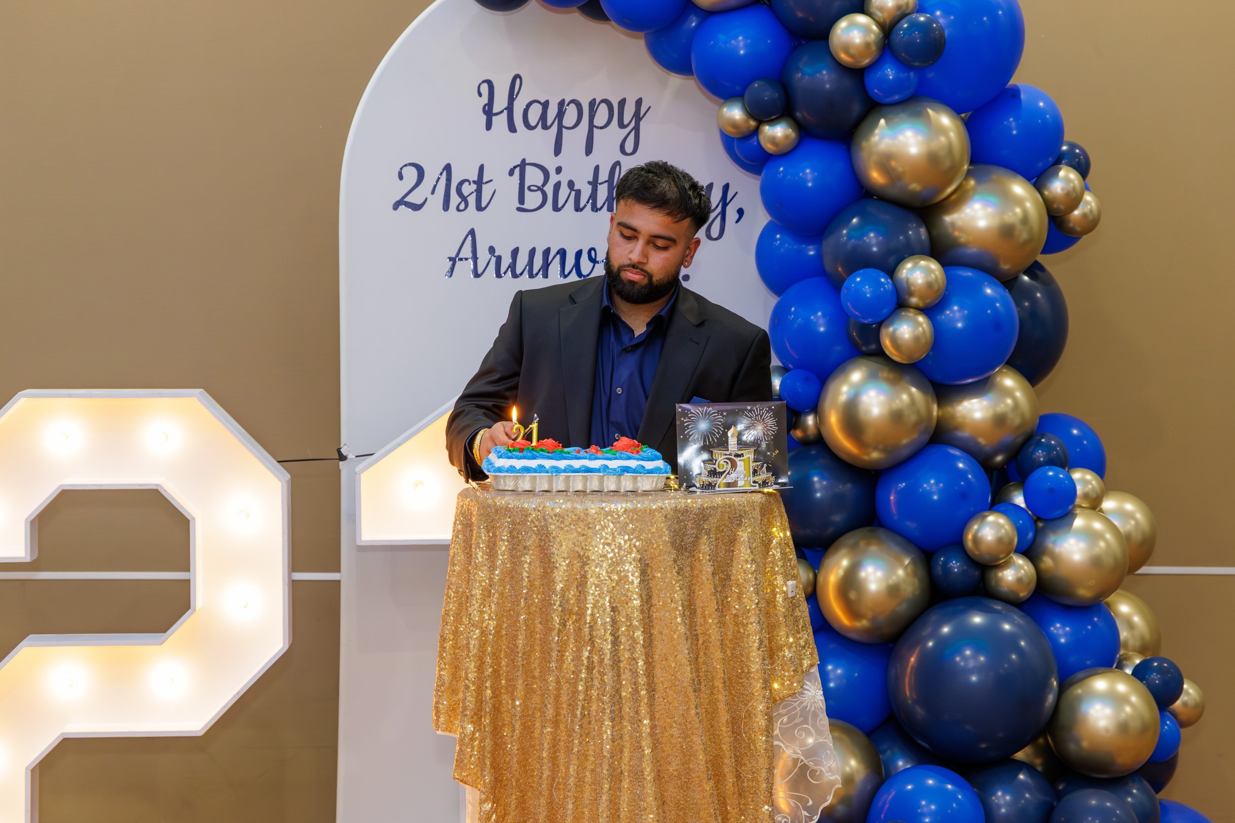 A man in a black suit and blue shirt is standing behind a table with a birthday cake, pink and blue decorations, in front of a backdrop with letters and balloons shaped as the number 21, celebrating his 21st birthday.