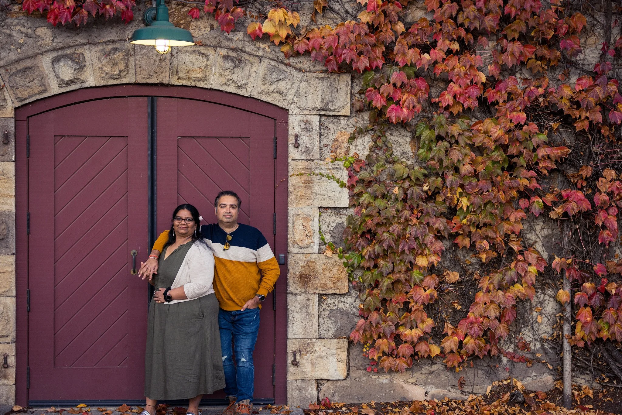 Two people standing in front of a maroon-colored, arched wooden door with autumn leaves on the ground and climbing ivy with red, orange, and green leaves on a stone wall.