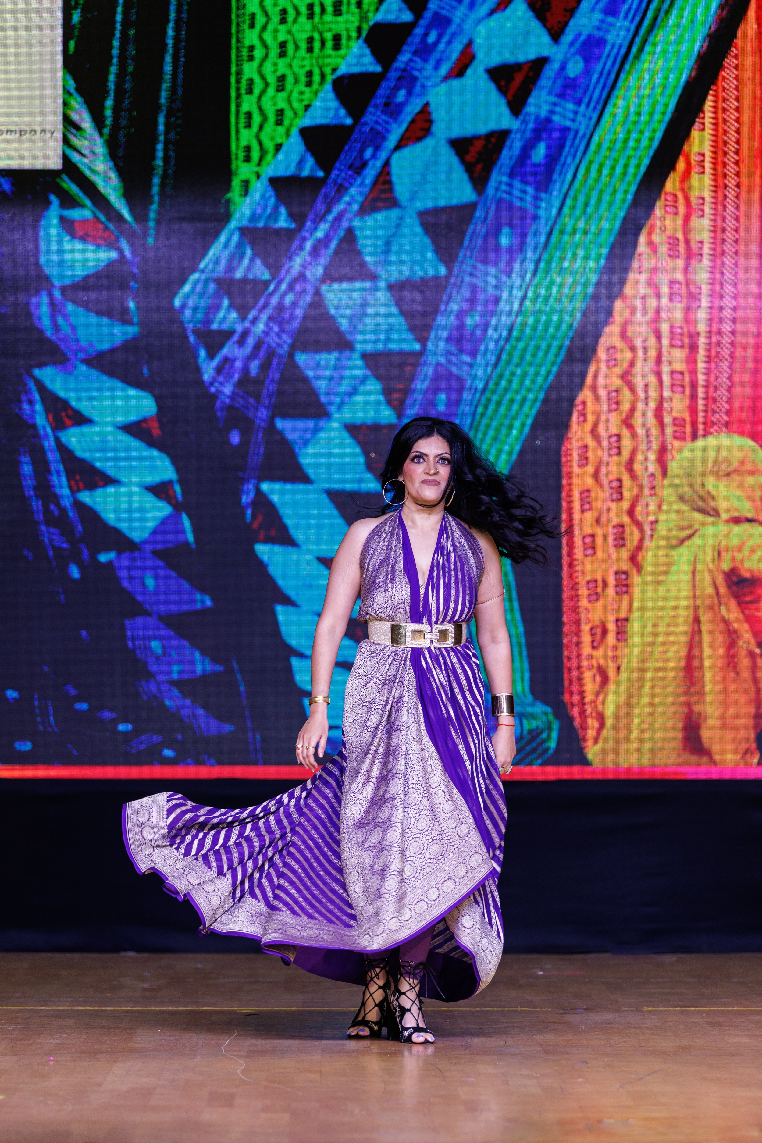 A woman walking on a runway wearing a long purple and white patterned dress with a beige belt, black lace-up high heels, and jewelry, against a colorful textured background.