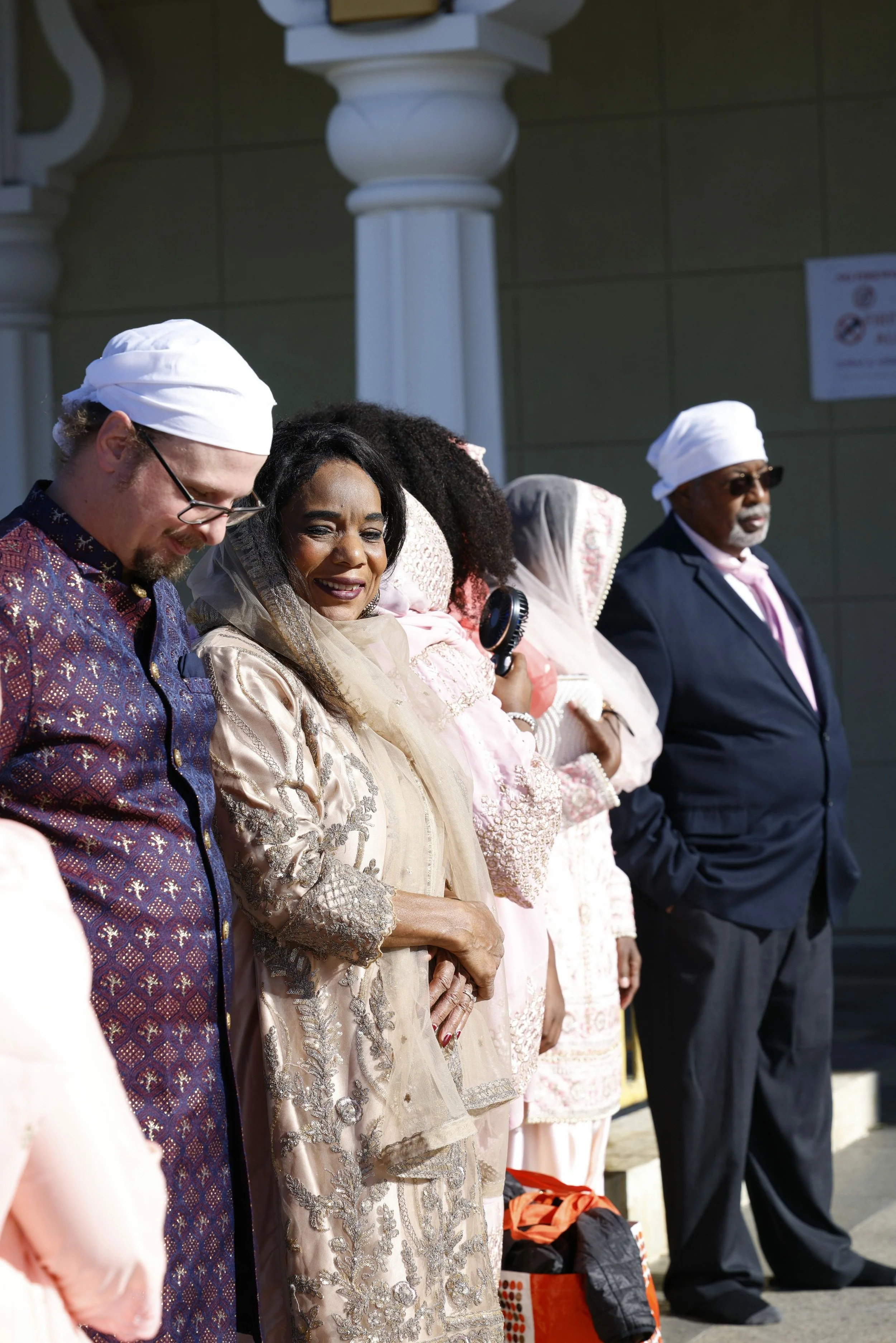 Group of people standing outdoors, dressed in traditional and formal attire, with some wearing turbans and head coverings, on a sunny day in front of a building with columns.