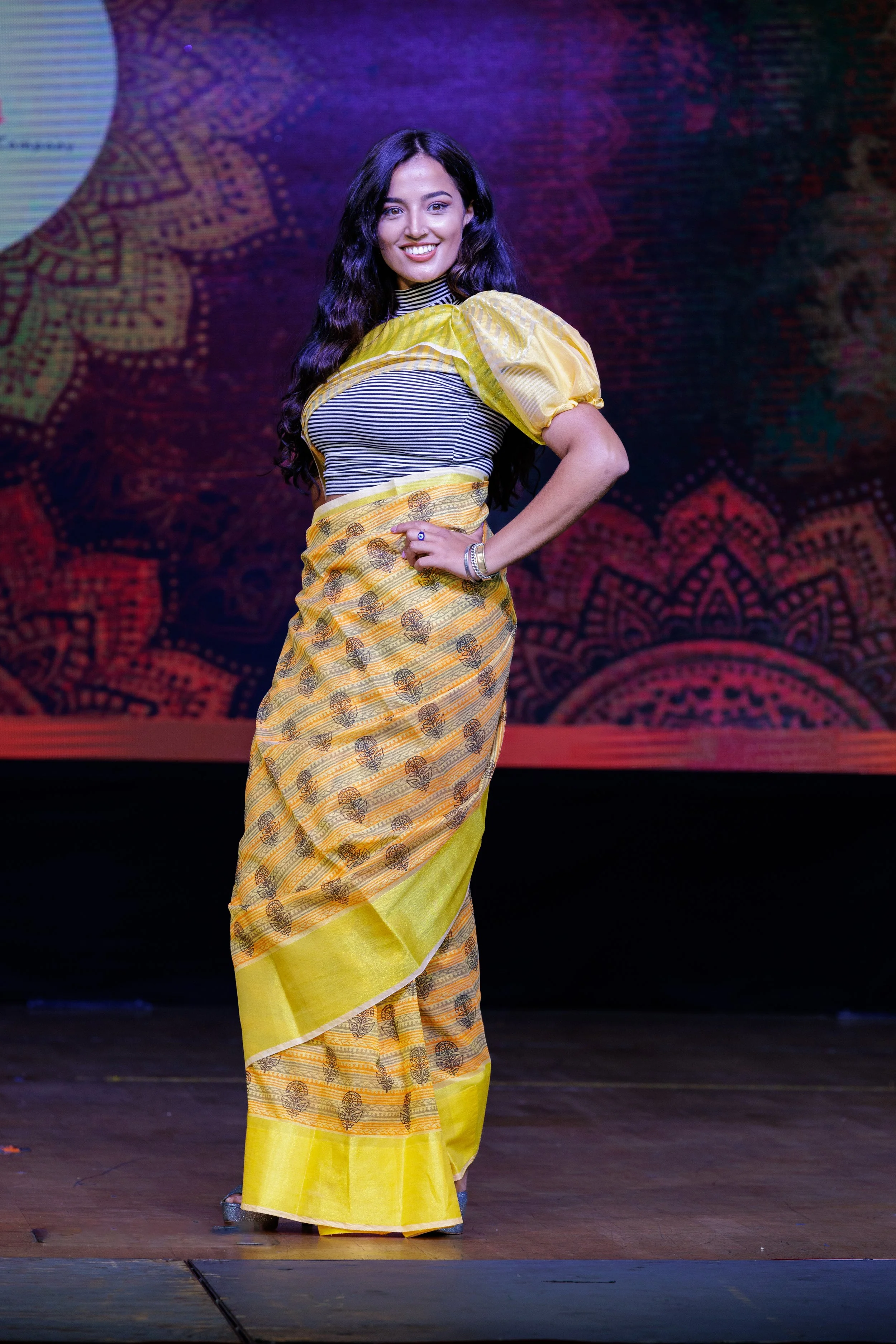 A woman standing on stage in a yellow and black saree, smiling at the camera, with a colorful, artistic background behind her.