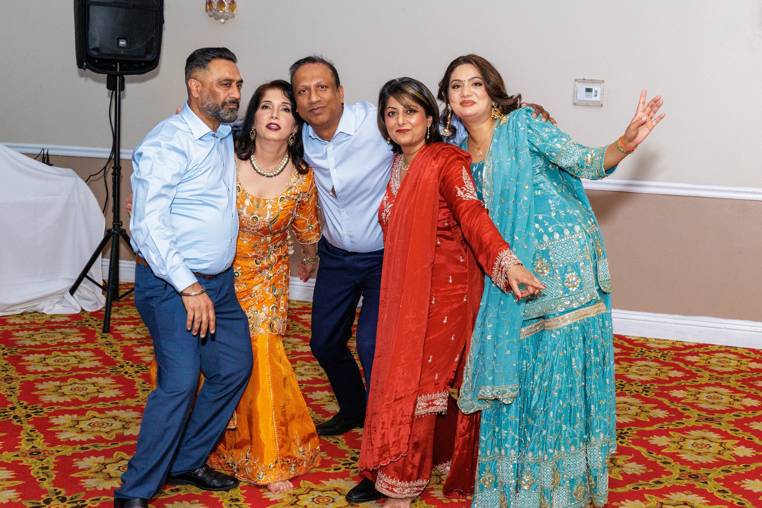 Six adults dressed in colorful traditional Indian clothing, standing together and posing for a photo at an indoor event with a red and gold patterned carpet.