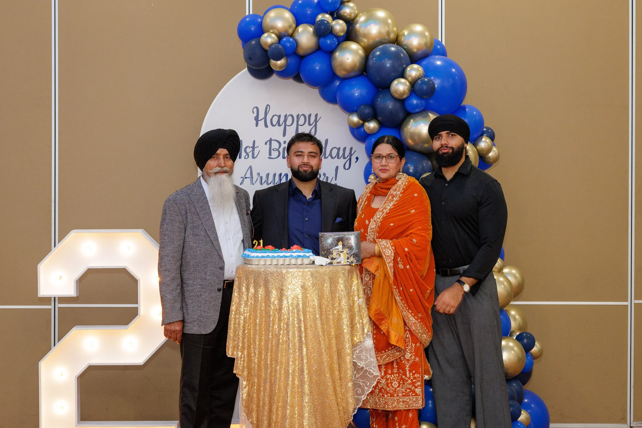 A family celebrating a birthday in front of a decorative backdrop with balloons and a sign that reads 'Happy 1st Birthday, Arunee!'. Four people are standing around a table with a birthday cake and a card, all dressed in formal attire.