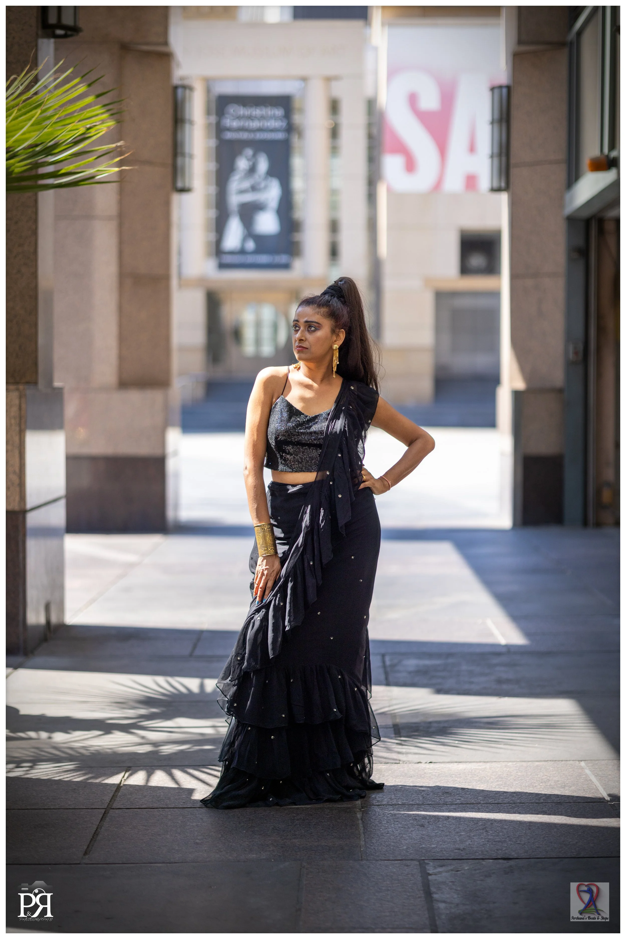 A woman with long dark hair in a high ponytail wearing a sparkly black top and long black ruffled skirt stands with one hand on her hip in an urban outdoor setting, with blurred city buildings and a sale sign in the background.