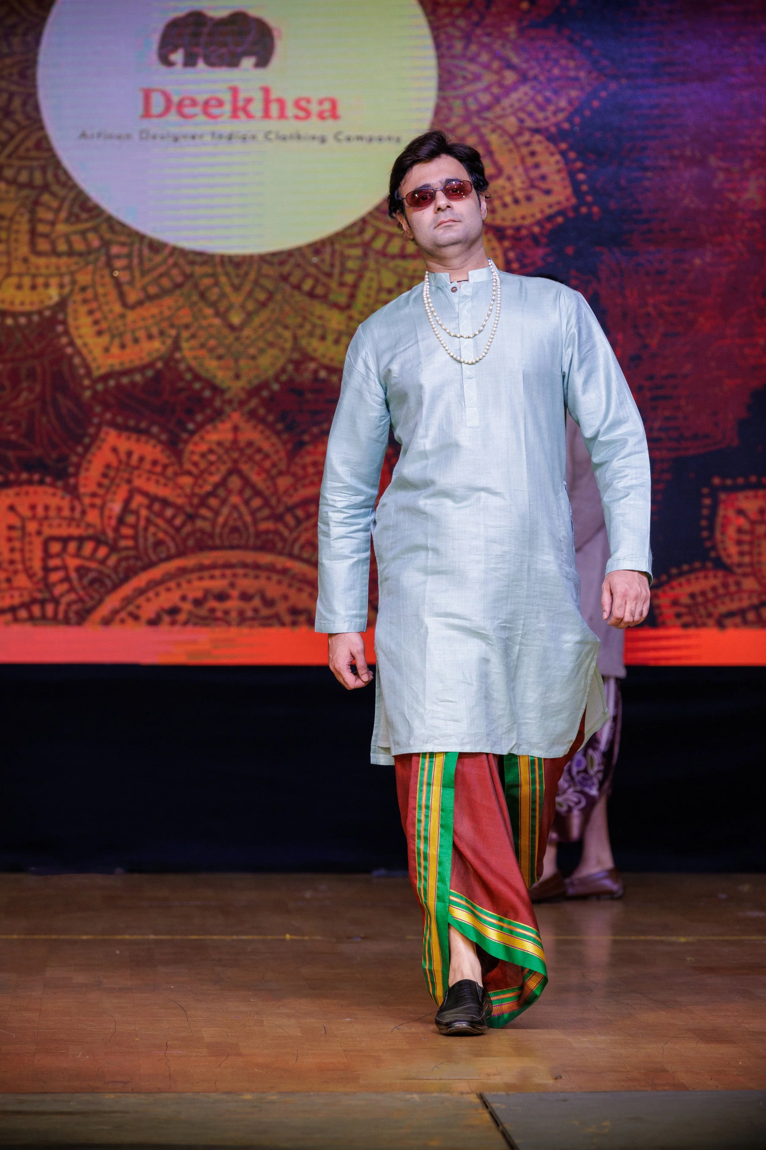 A man walking on a runway wearing traditional South Asian attire, including a light-colored kurta, striped pants with green, yellow, and red accents, dark shoes, and sunglasses, with a large logo for 'Deeksha' in the background.