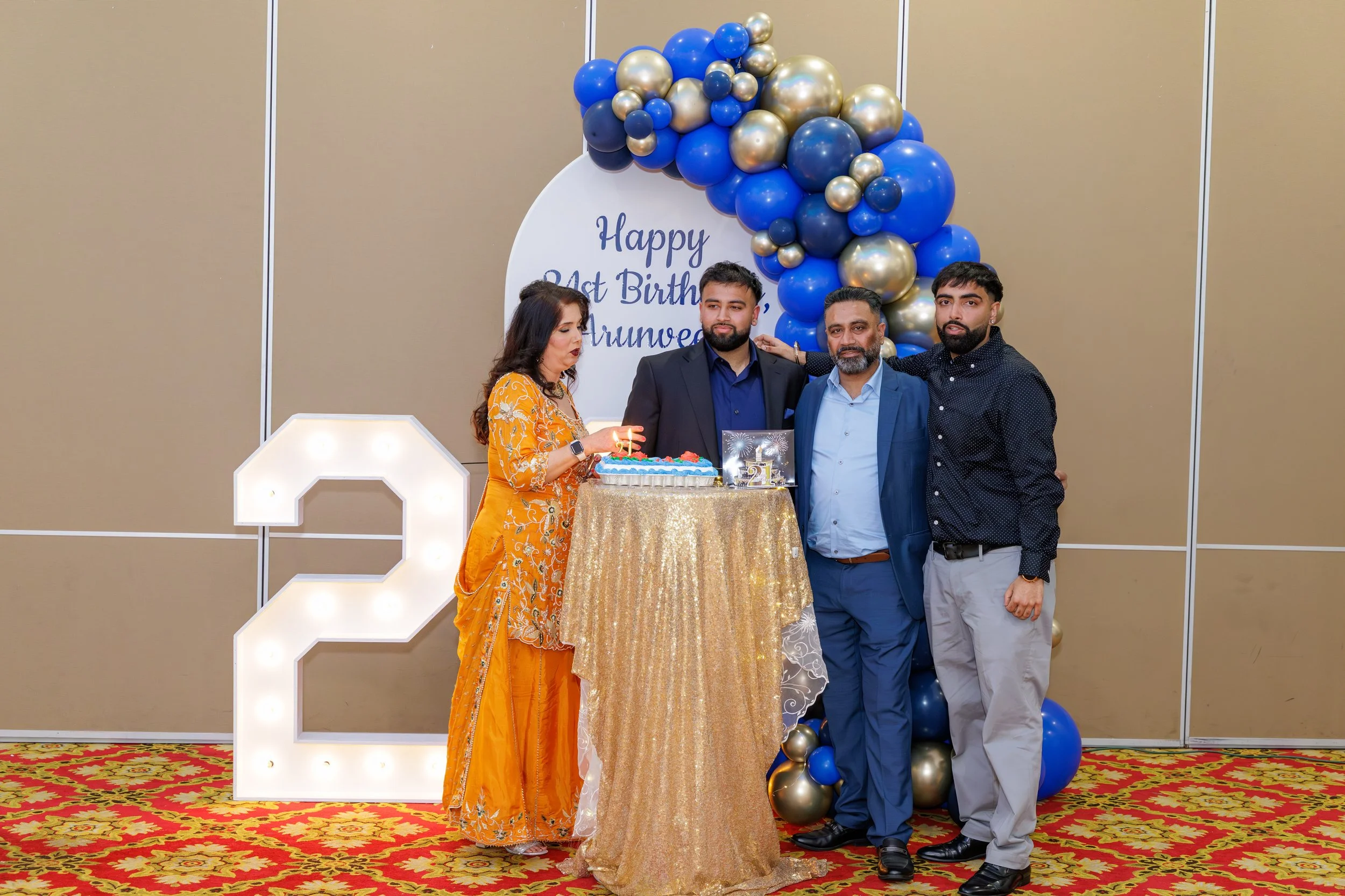 A family celebrating a 21st birthday at an indoor party with a gold and blue balloon arch, a large illuminated number 2, and a decorated table with a birthday cake. There are four people in formal attire, a woman in an orange saree, and three men in 