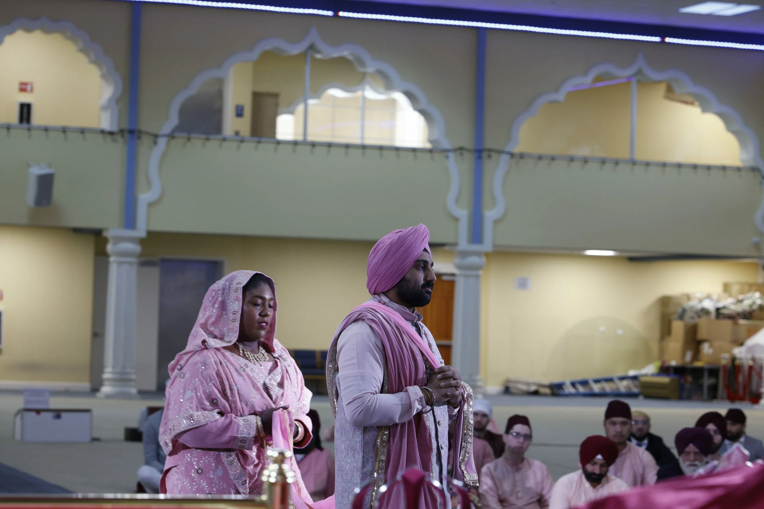 A couple dressed in traditional Indian wedding attire, with the groom wearing a pink turban and the bride in a pink outfit with a head covering, participating in a ceremony inside a decorated hall filled with seated guests.