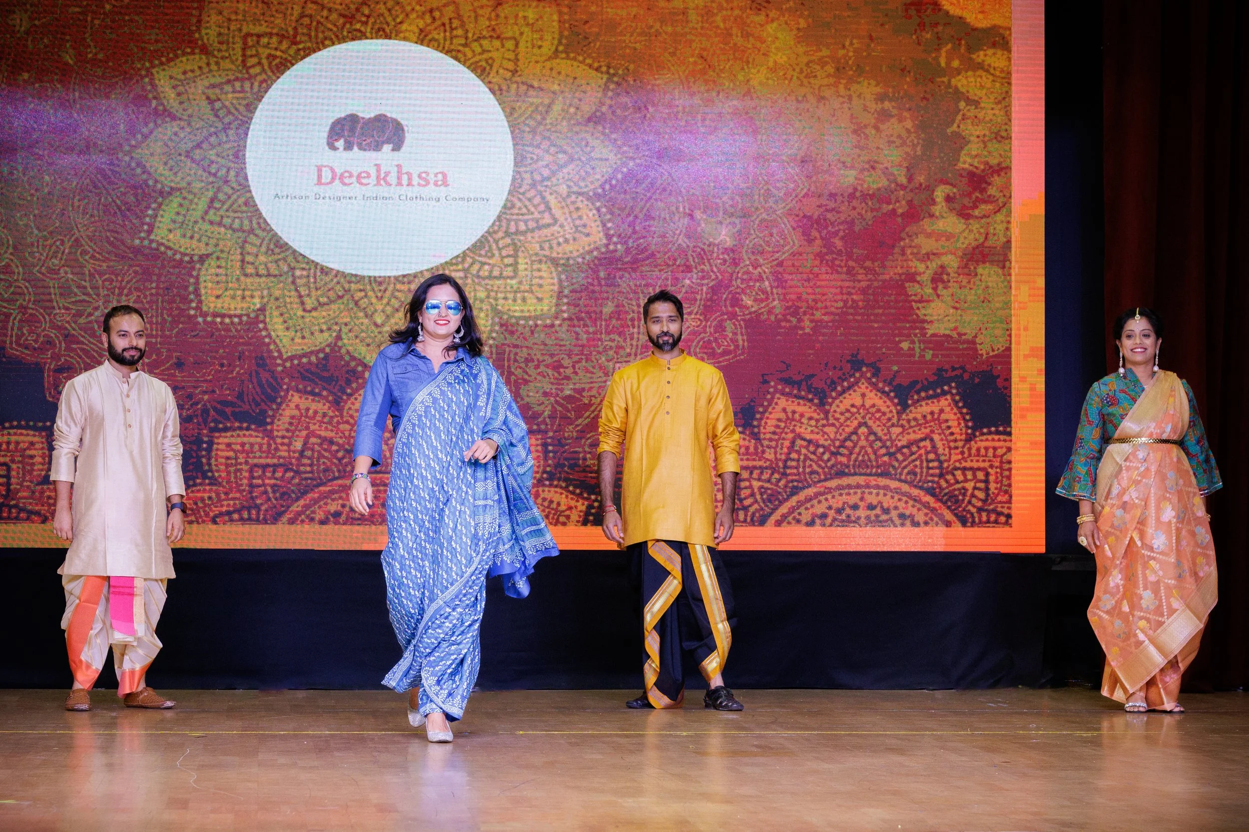 Four people on stage dressed in traditional Indian attire, standing in front of a colorful backdrop displaying a logo and the word 'Deekhsa'.