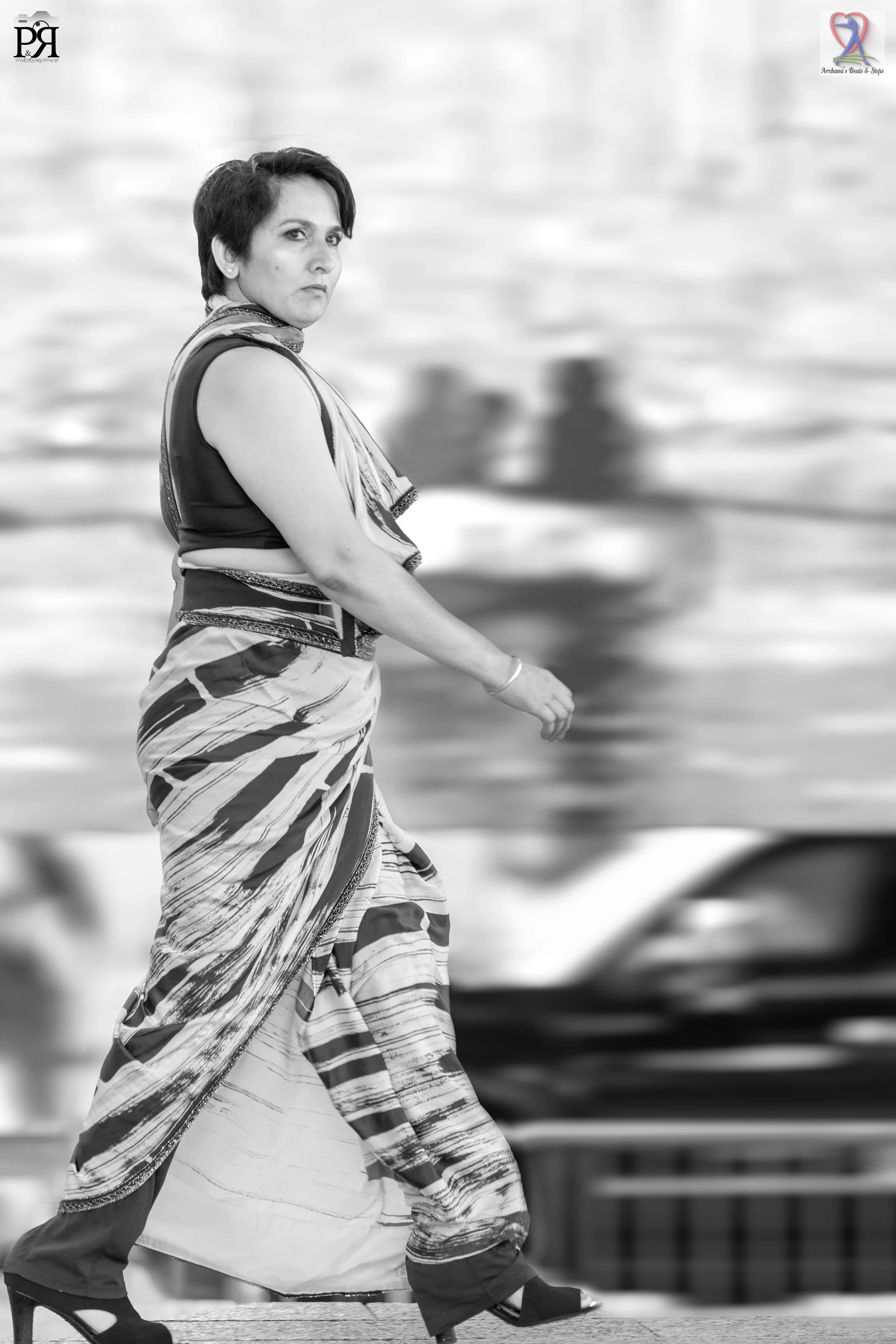 A black and white photo of a woman in traditional Indian attire walking along a waterfront promenade with boats in the background.