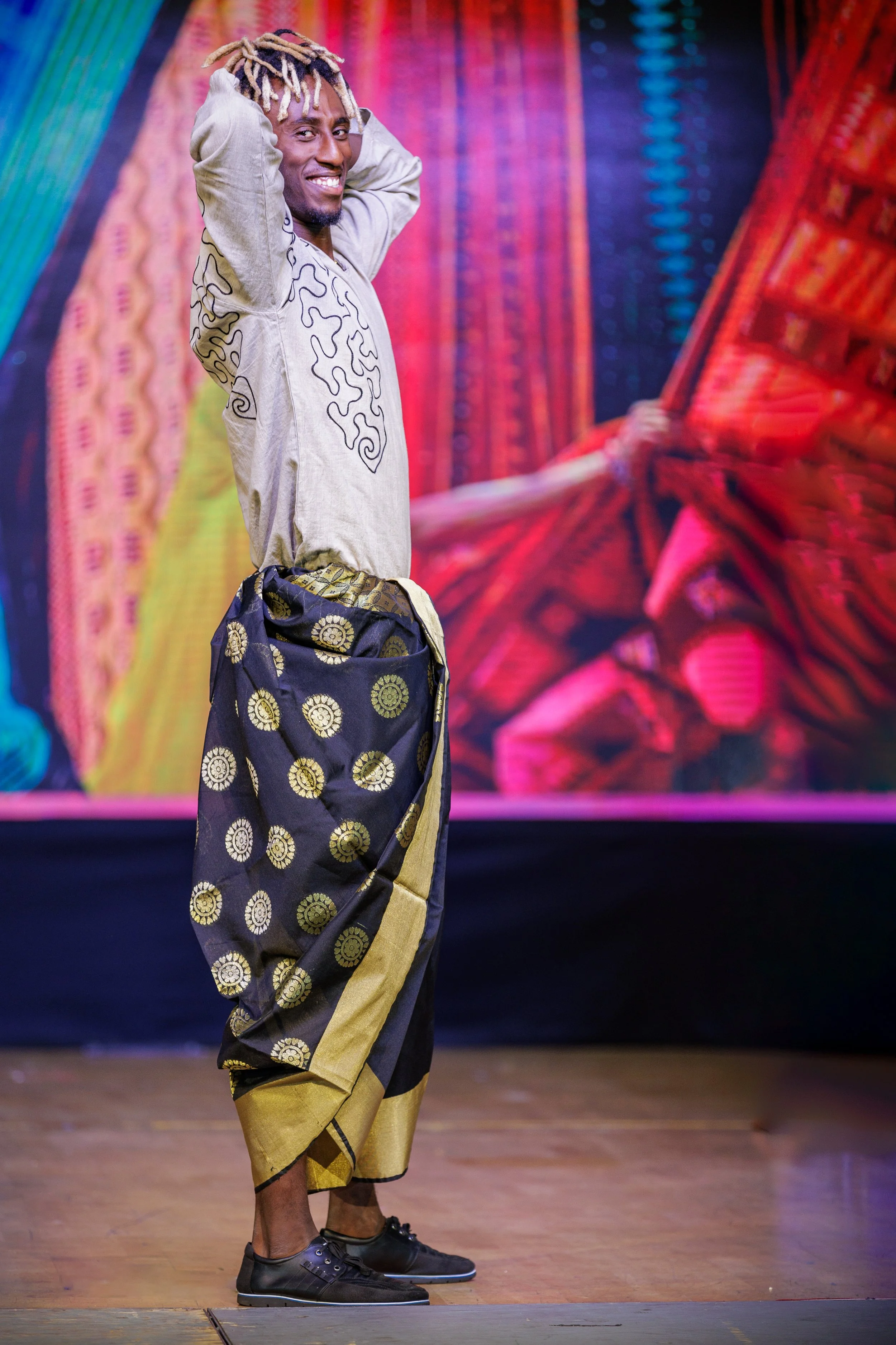 A man with dreadlocks smiling and holding his hands behind his head, standing on a stage with a colorful backdrop. He is dressed in a loose beige shirt with black designs, patterned wide-leg pants, and black shoes.