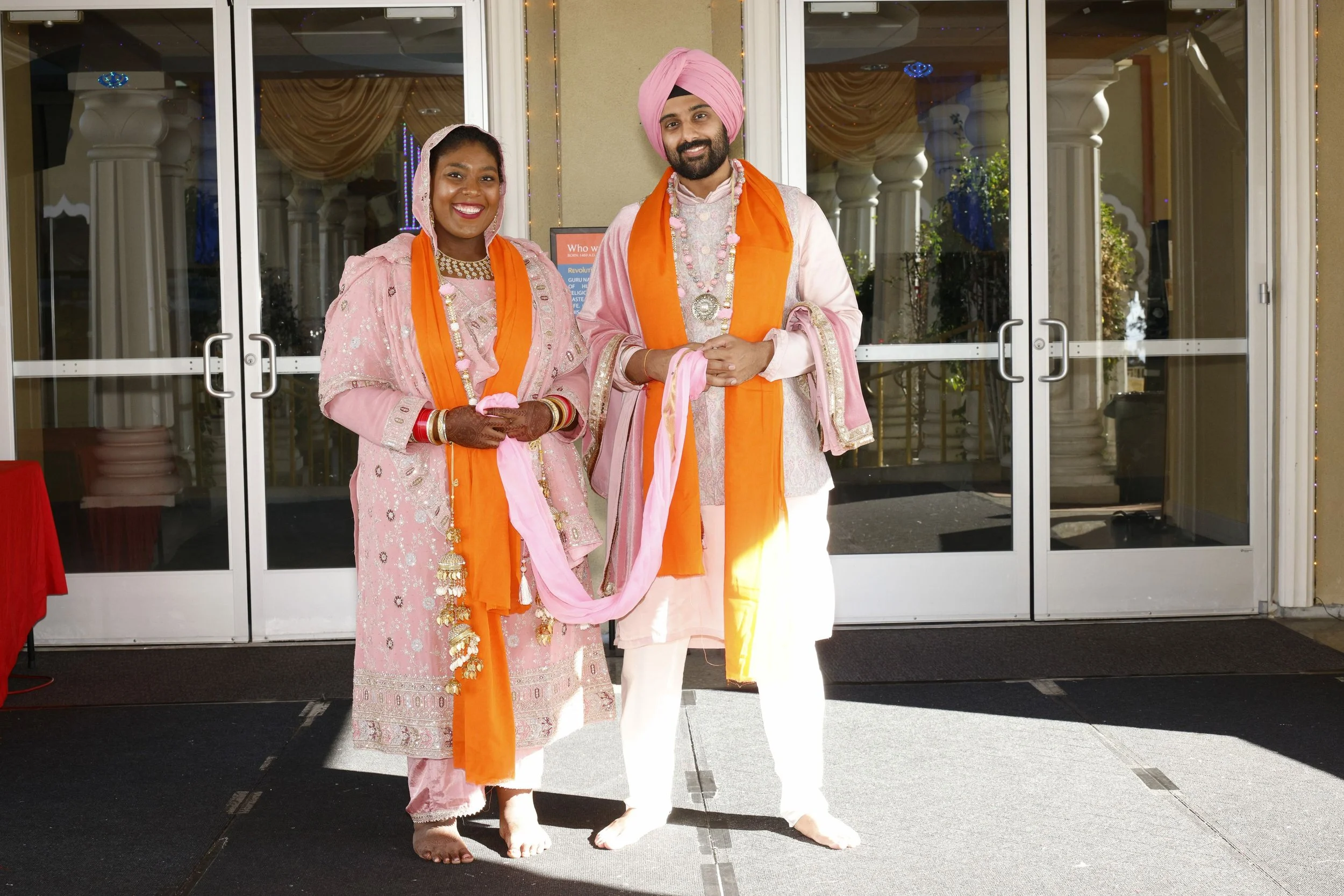 A man and woman dressed in traditional Indian wedding attire, standing side by side indoors in front of glass doors, smiling at the camera, with orange scarves and pink clothing.