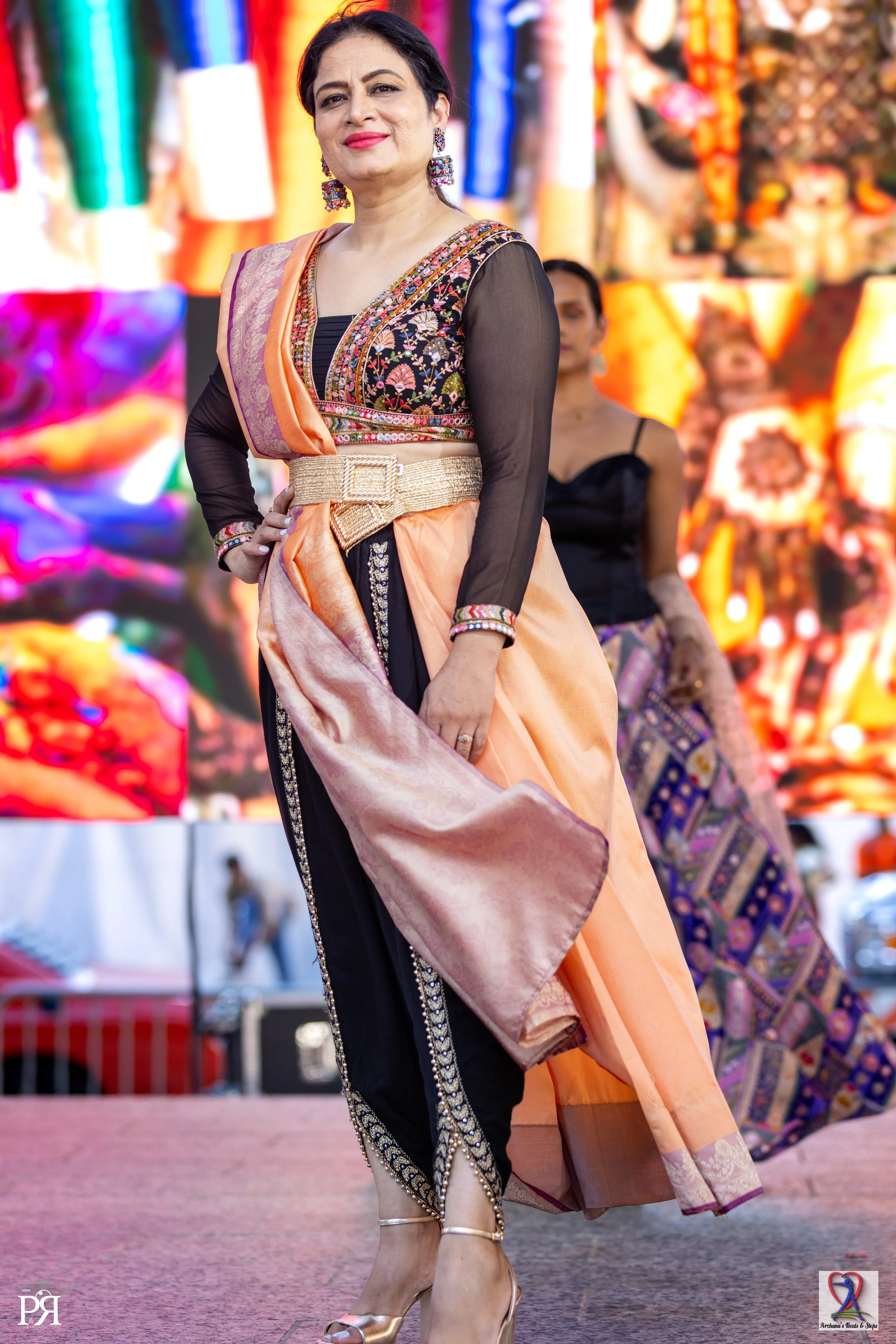 A woman dressed in colorful traditional Indian attire, standing confidently on a stage during a cultural event with a vibrant backdrop.