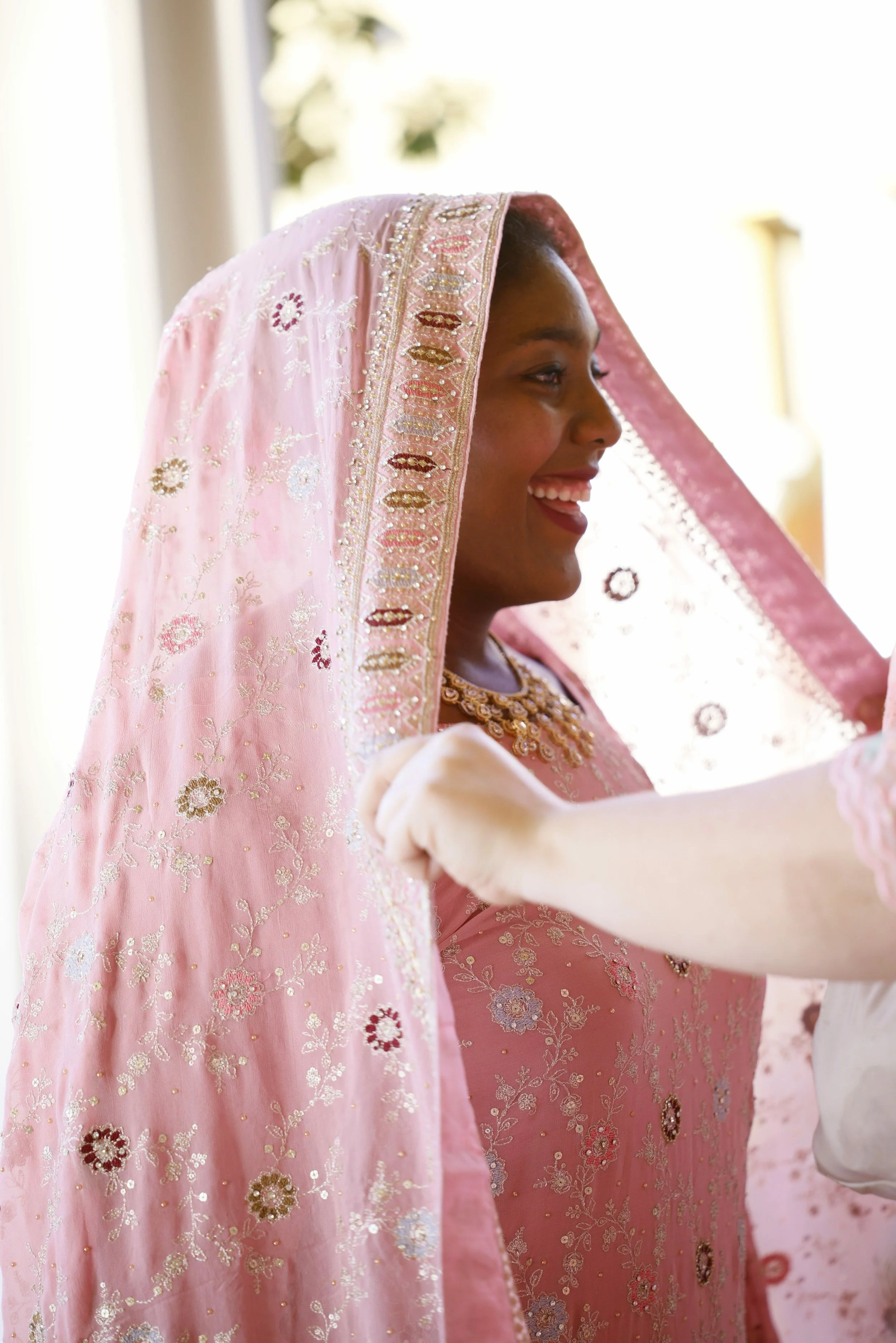A woman dressed in a pink, embroidered traditional attire, smiling as someone puts a matching pink head covering on her head.