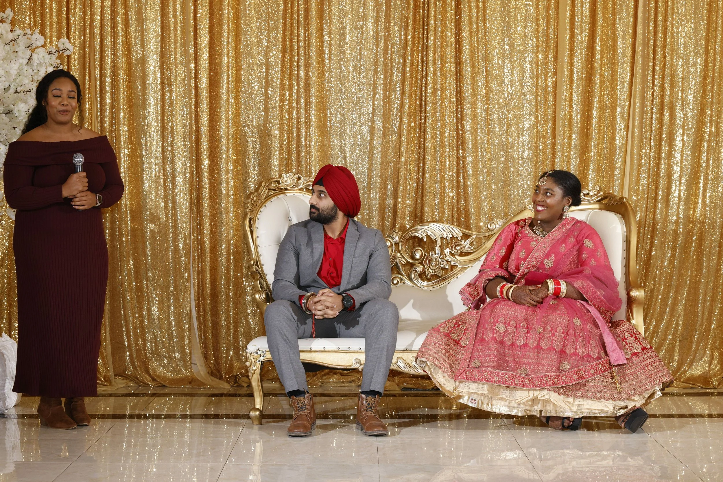 A woman in a maroon dress stands holding a microphone, talking to a man in a gray suit with a red turban and a woman in a pink traditional dress seated on an ornate gold and white sofa, against a golden shimmering curtain backdrop at a celebration ev