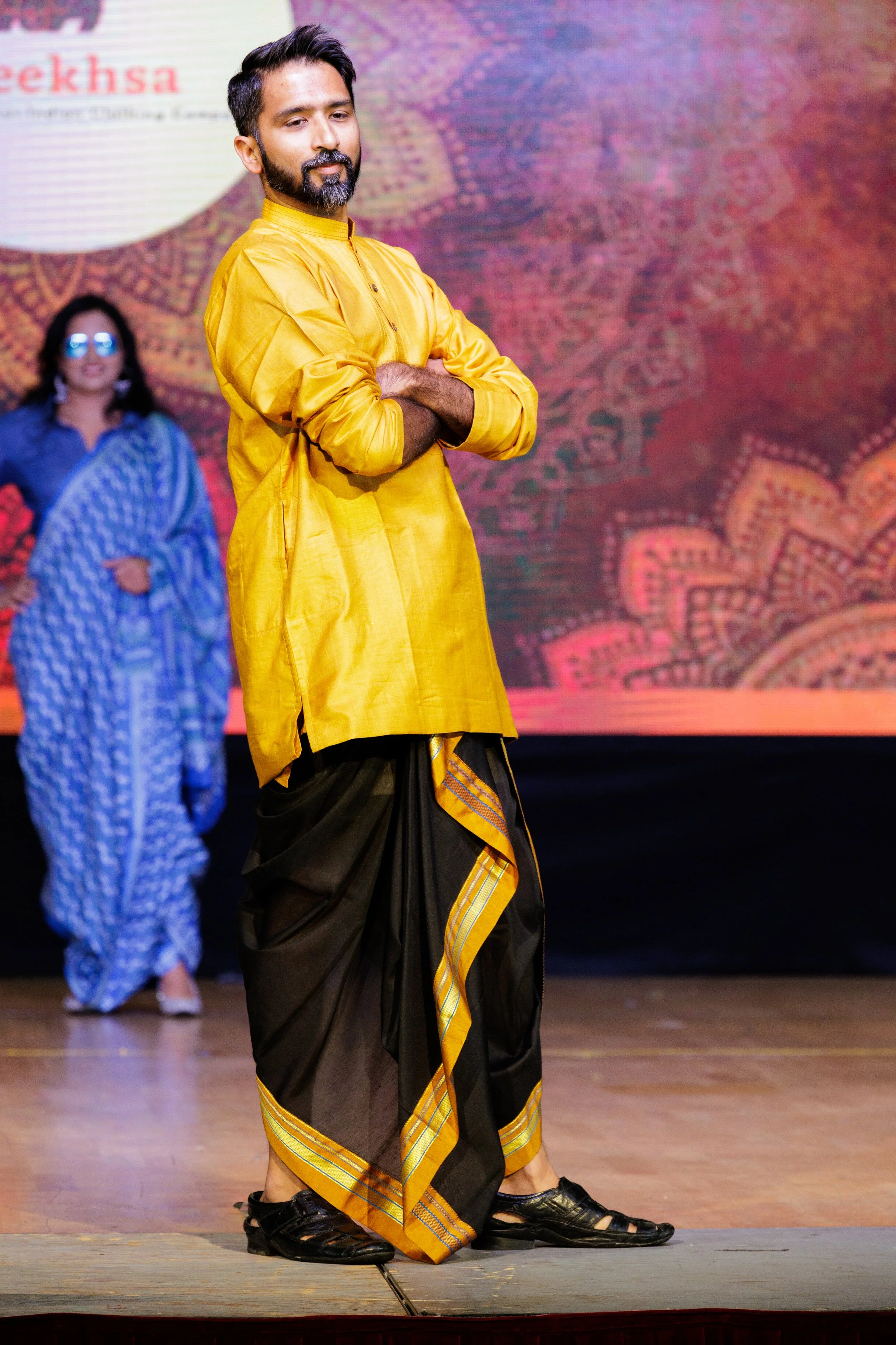 Man wearing a yellow kurta with black and yellow dhoti, standing on stage with arms crossed, in front of a woman in a blue sari with glasses, and a backdrop with colorful patterns and text.