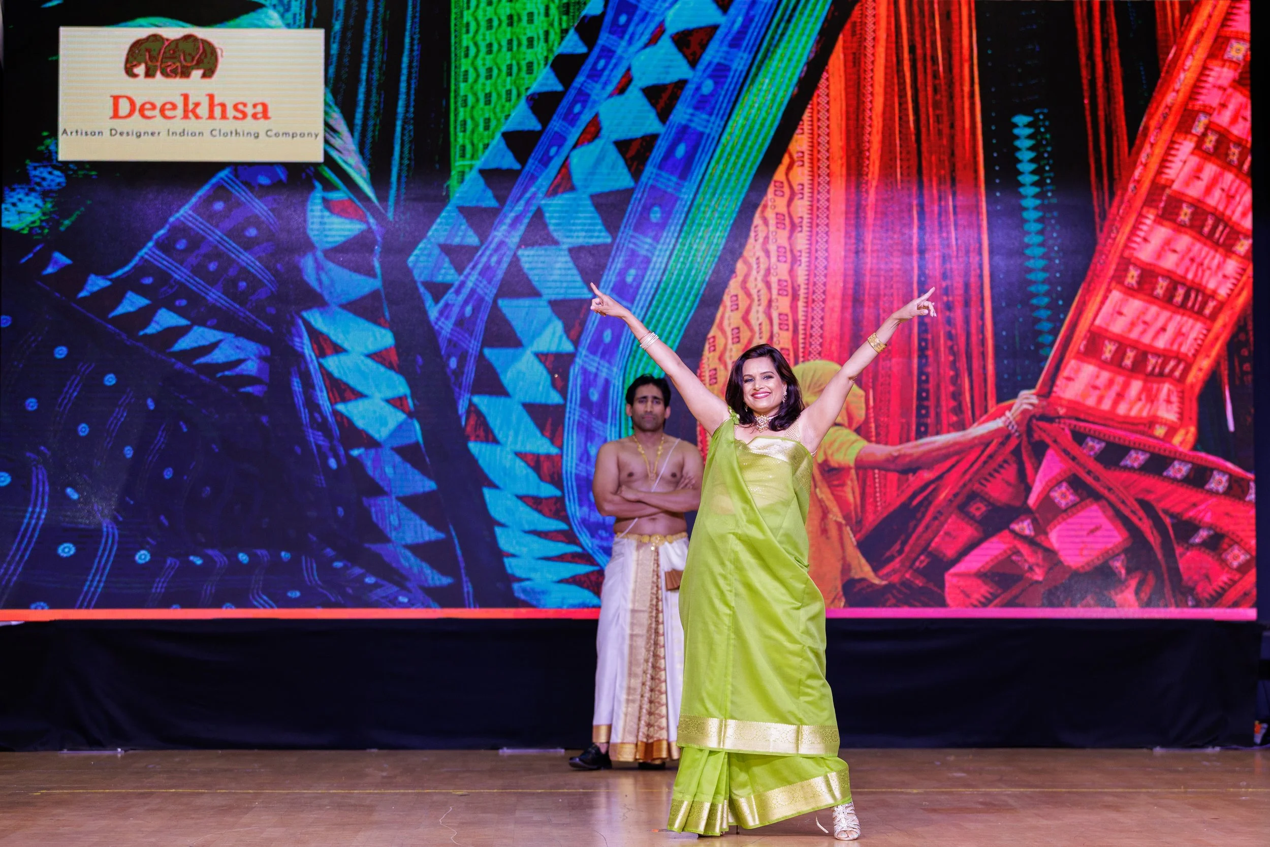A woman in a bright green traditional dress dancing on stage with her arms raised, smiling, with a man dressed in traditional attire standing behind her. The background features colorful, intricate cultural patterns and a logo in the top left corner 