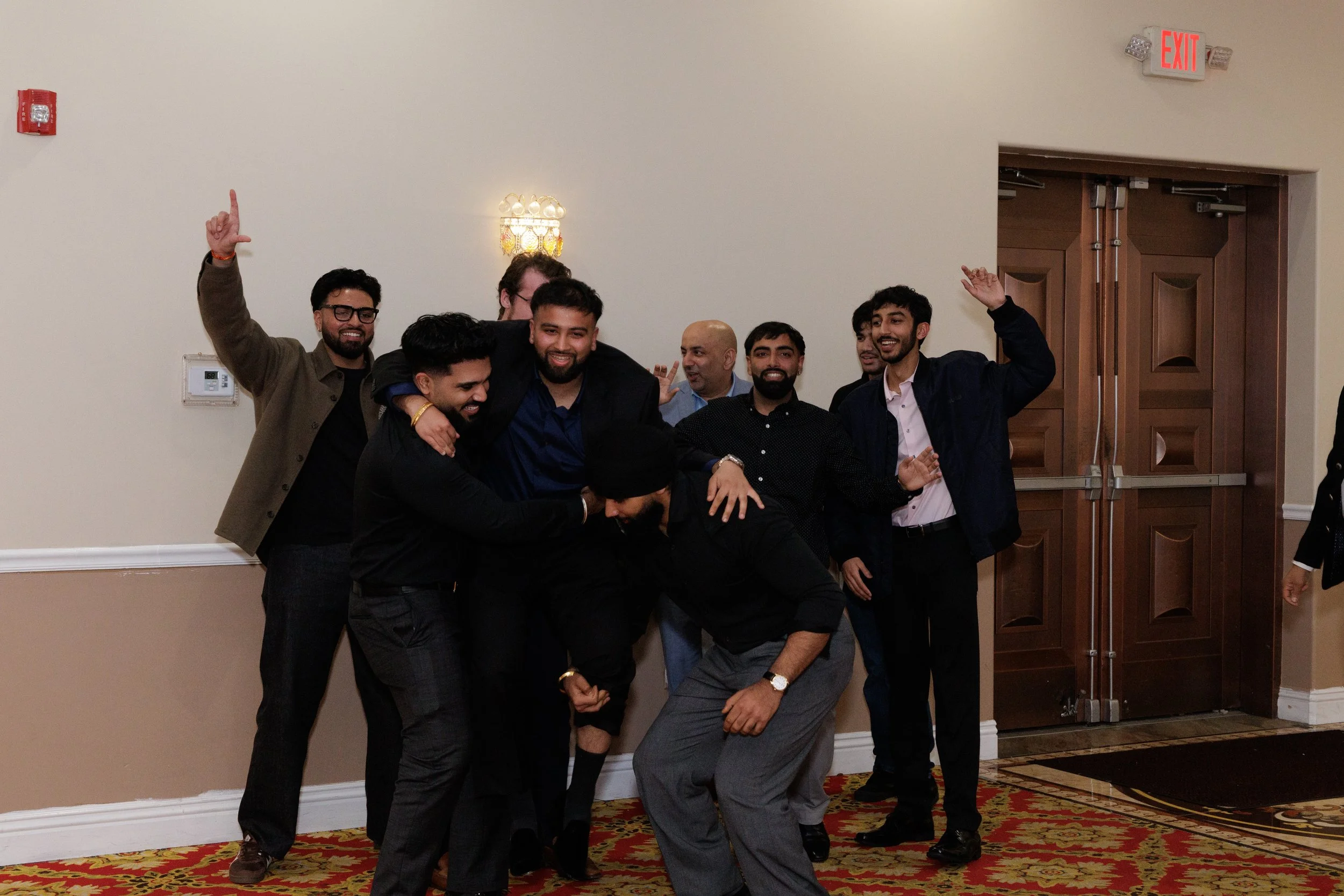 Group of men celebrating and smiling at an indoor event, standing in front of a wooden door and beige wall with an exit sign above.