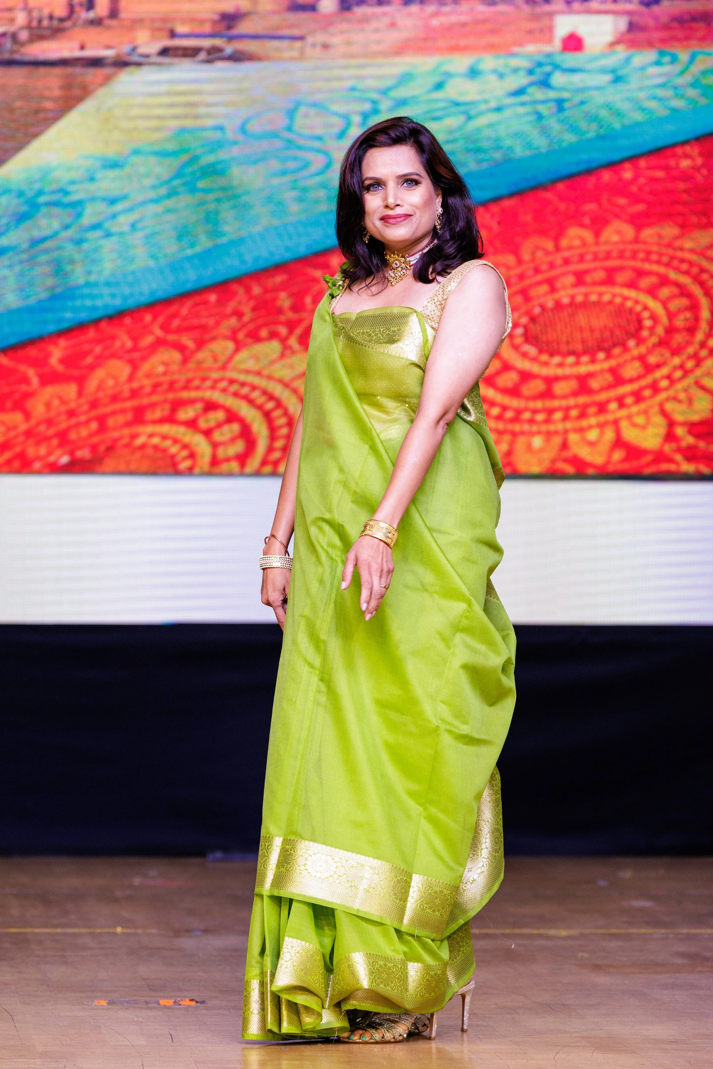 Woman wearing a lime green saree with gold border, standing on a stage with colorful digital background.