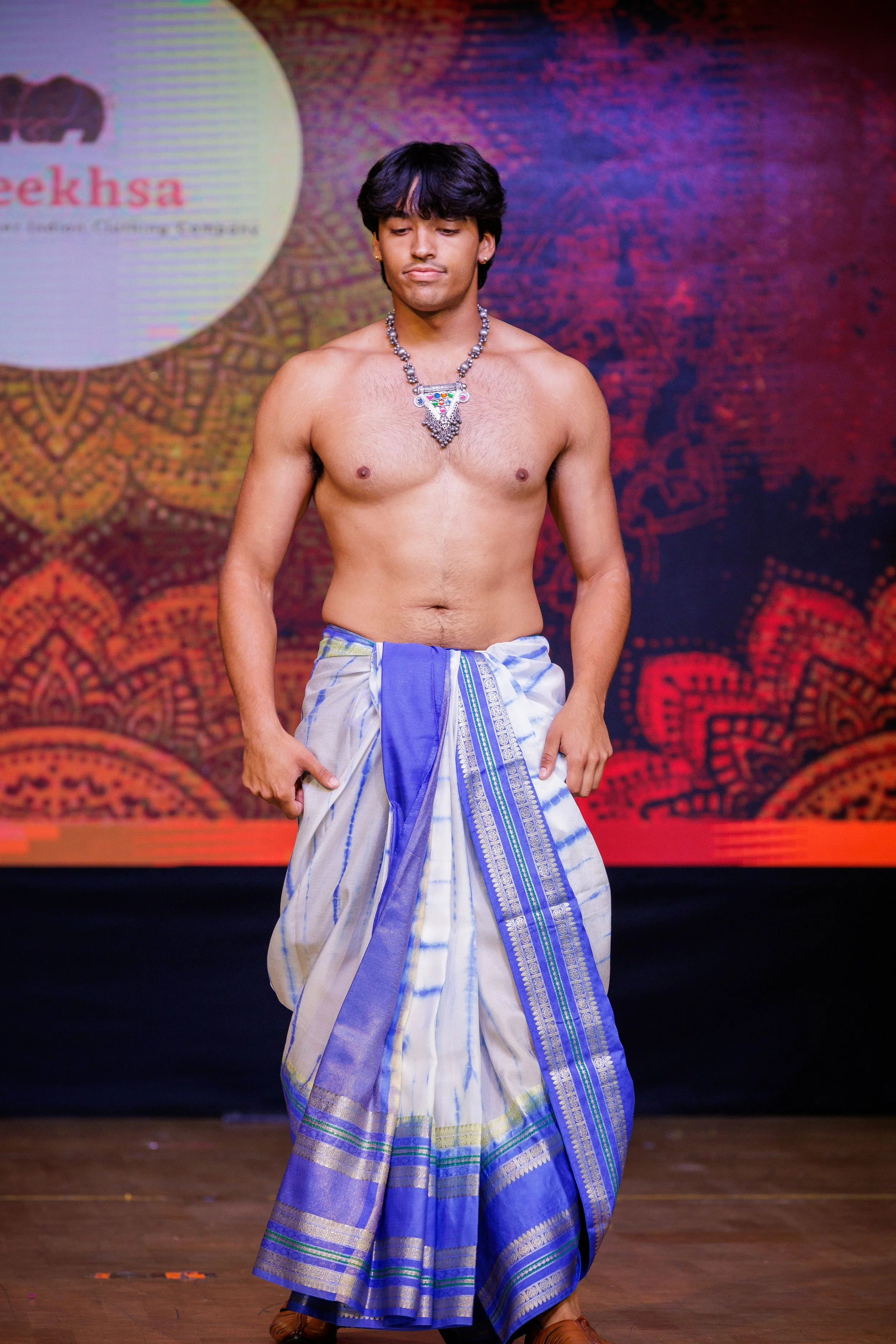 A young male model walking on stage wearing traditional Indian dhoti with a tribal necklace, with a colorful background and a logo visible behind him.