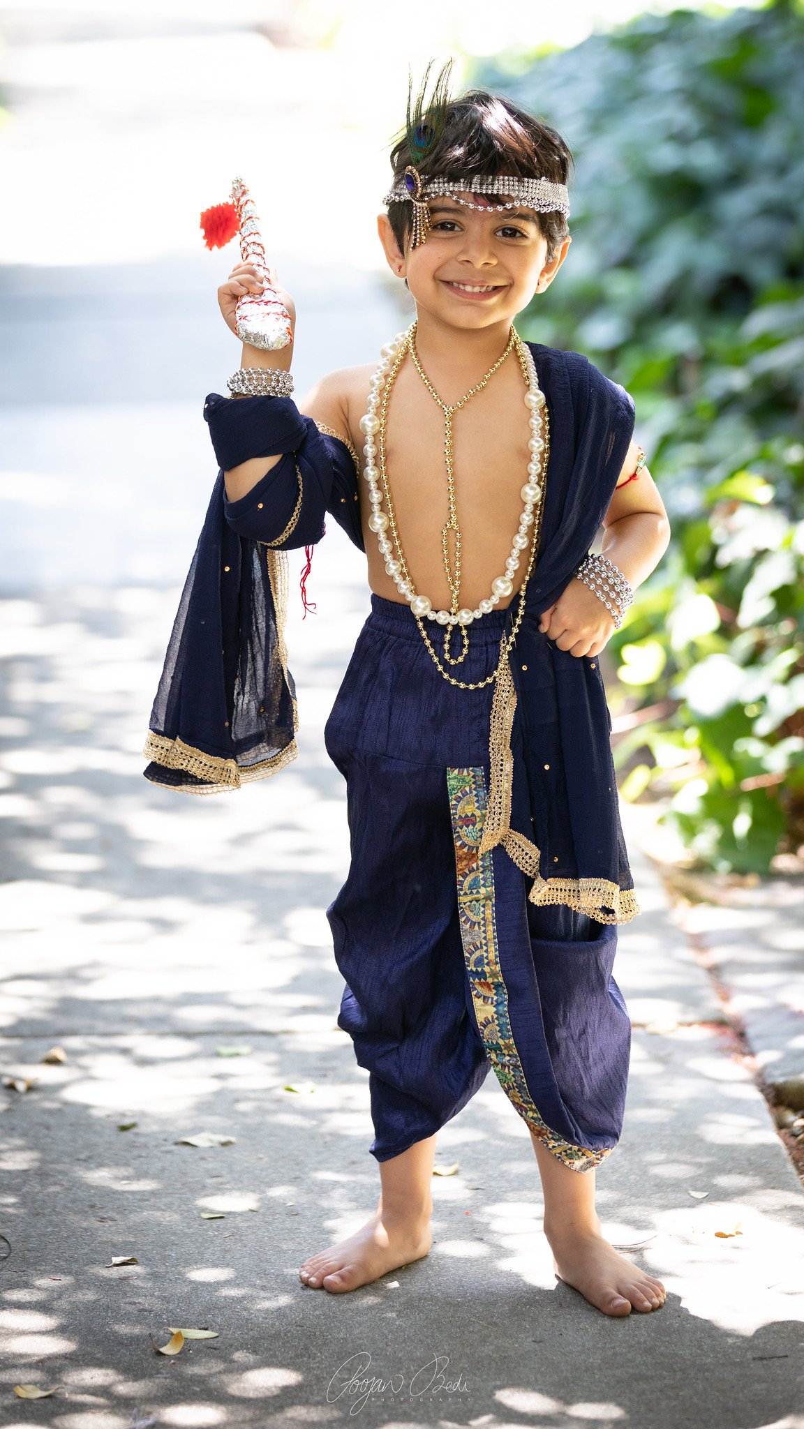 A young boy dressed as a traditional Indian bride, wearing blue saree with gold embroidery, multiple pearl and gold necklaces, bracelets, and a headpiece, holding a decorative item, smiling outdoors.