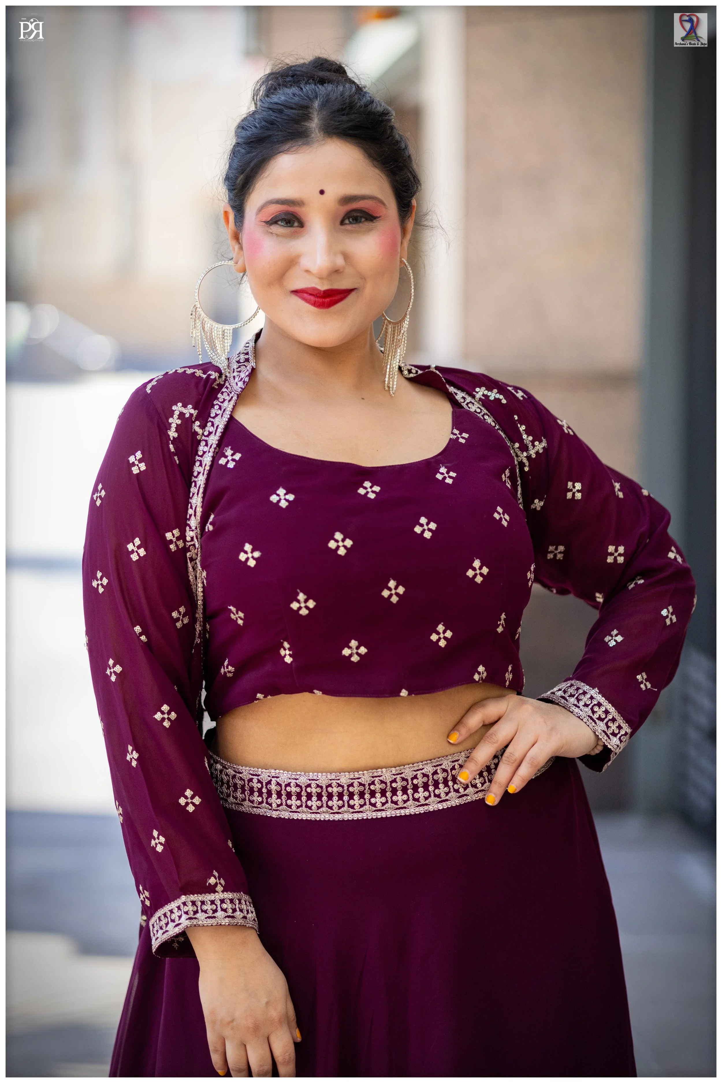 Woman dressed in a deep purple traditional Indian outfit with silver embroidery, wearing large silver hoop earrings, and bright makeup, standing outdoors in front of buildings.