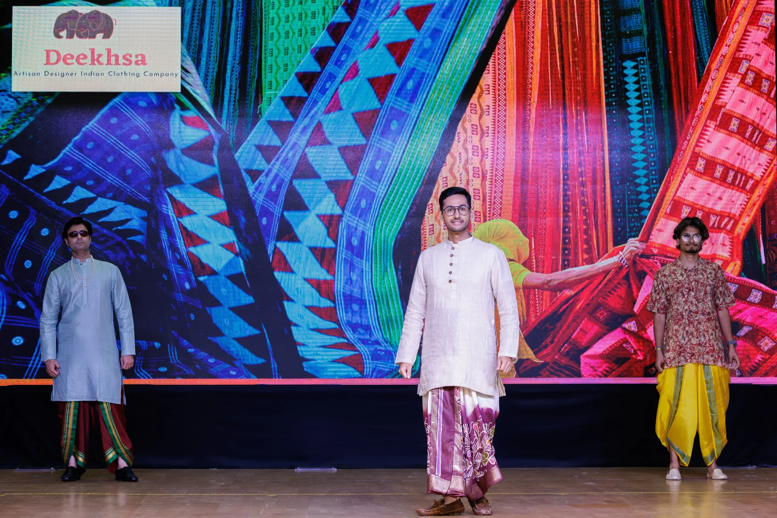 Three male models on stage wearing traditional Indian clothing, with colorful woven fabrics in the background showcasing various patterns, at a fashion event for Deeksha, an Indian clothing company.