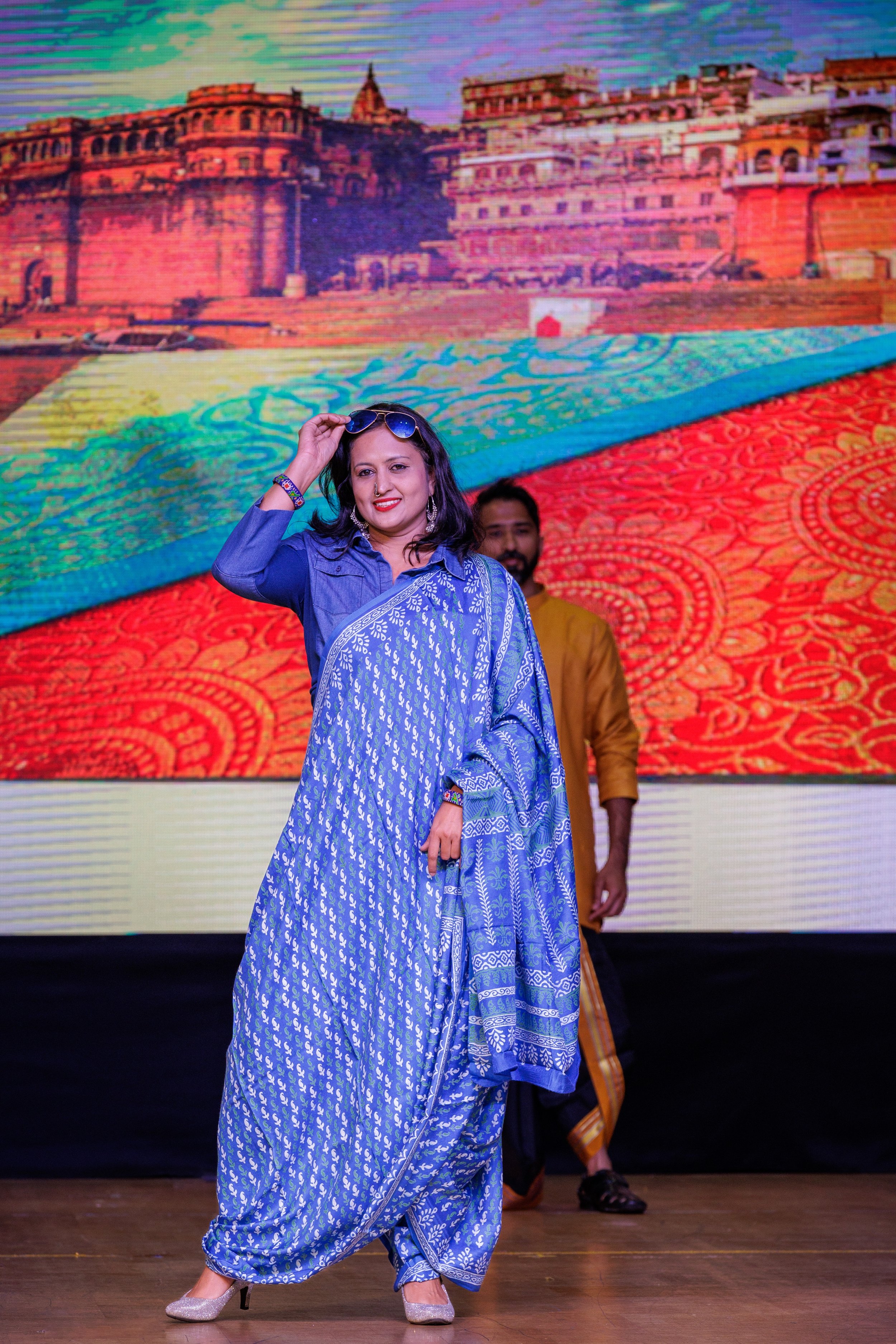 A woman in a blue saree posing on stage with a colorful background of an Indian palace and pattern. She is smiling and holding sunglasses, with a man in traditional Indian attire standing behind her.