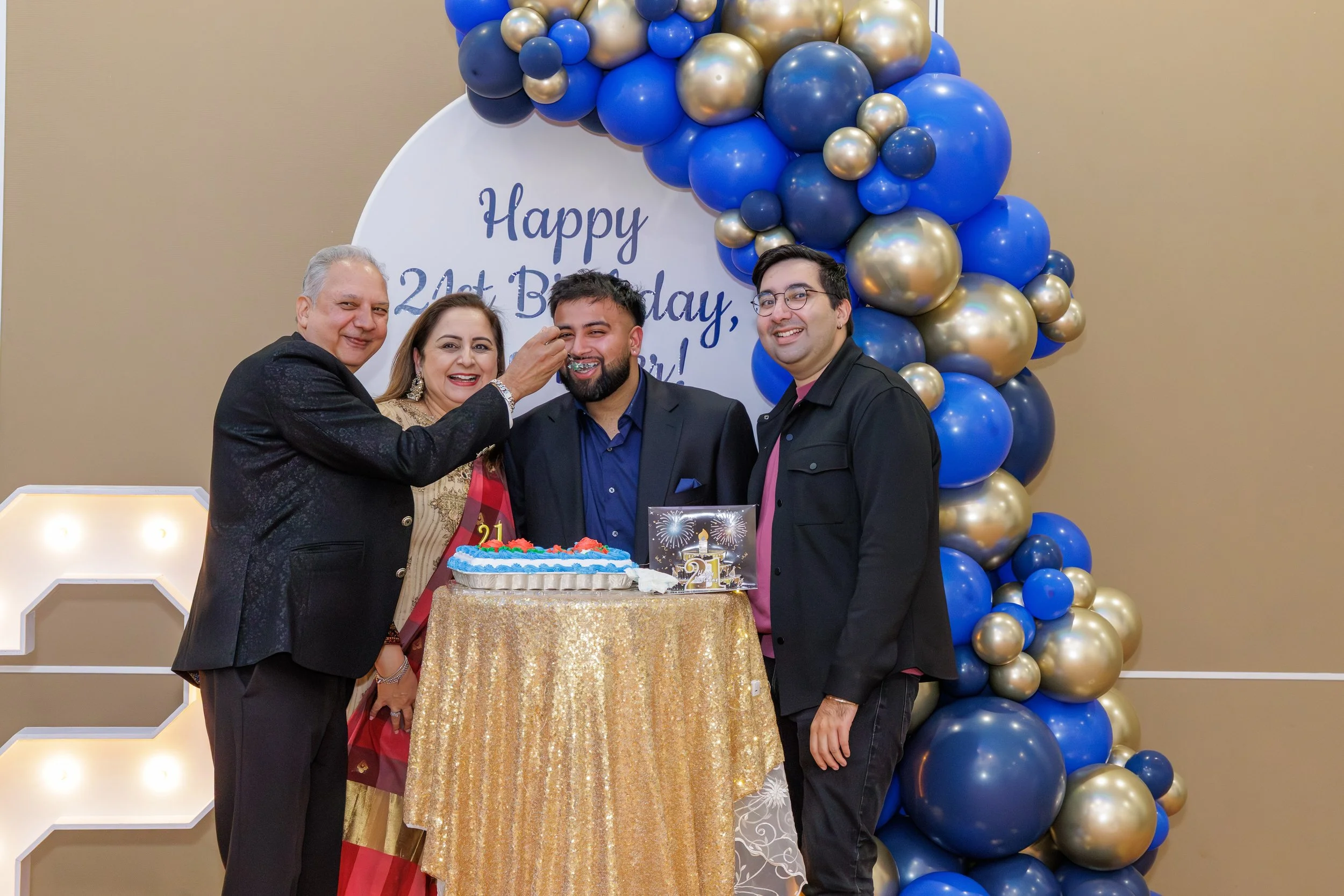 Four people celebrating a birthday together, with a decorated backdrop and a birthday cake on a golden table. One person is feeding the birthday celebrant, who is smiling with a cake piece in his mouth. The backdrop has a sign that says 'Happy 21st B