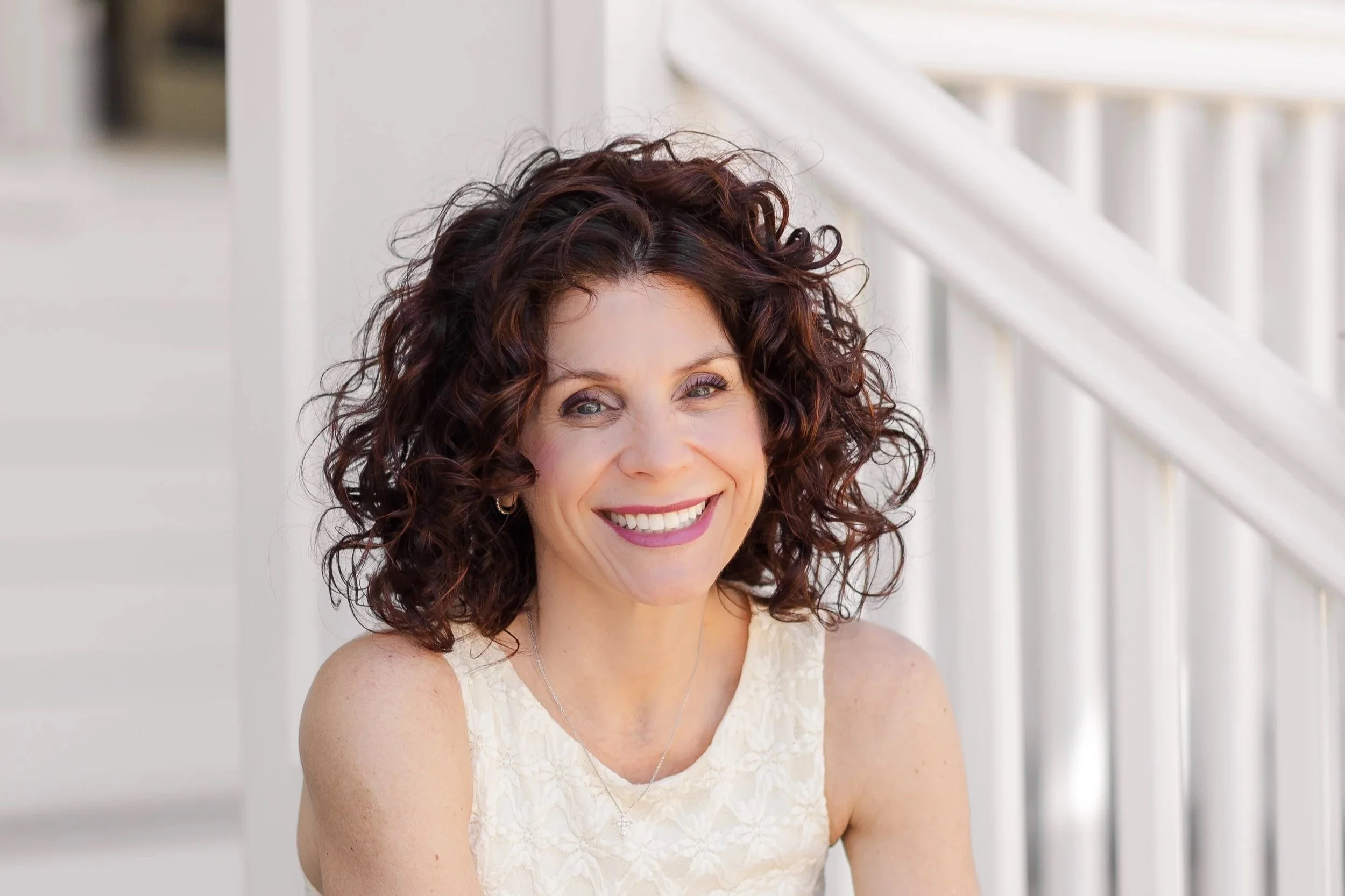A woman with curly brown hair and blue eyes smiling, wearing a cream-colored lace dress, standing in front of a white railing on a porch or balcony.