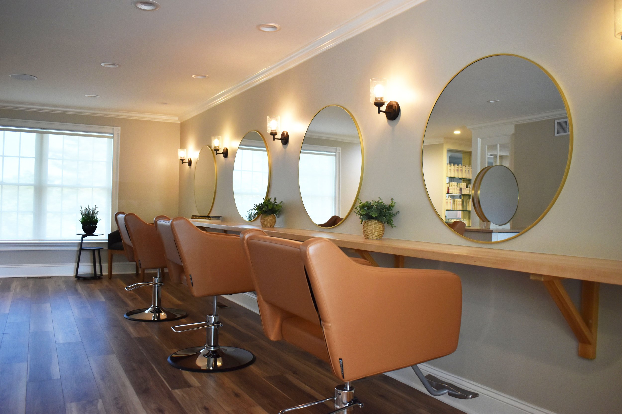 Beauty salon interior with a long mirror, pink salon chairs, wall-mounted lights, a window with white blinds, and small plants on the counter.